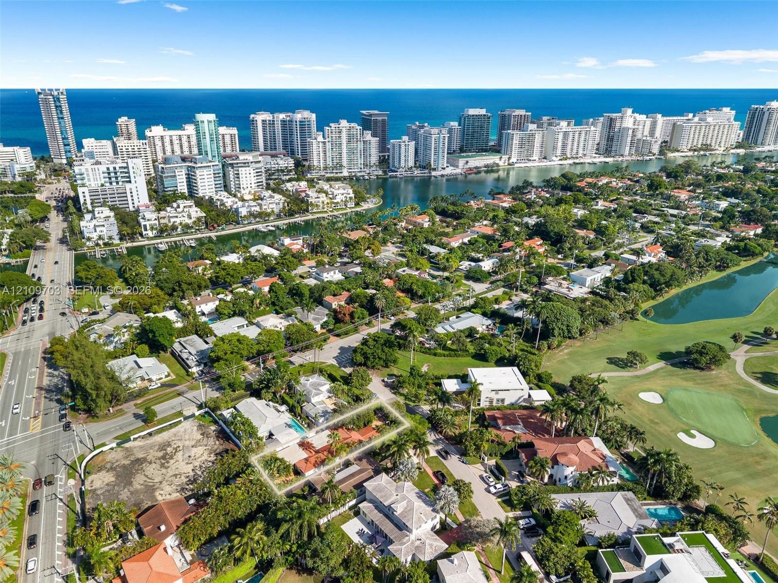 Aerial perspective featuring a single-story residence with a clay tile roof, a swimming pool, and mature palm trees, positioned in a coastal setting with proximity to a golf course and urban skyline
