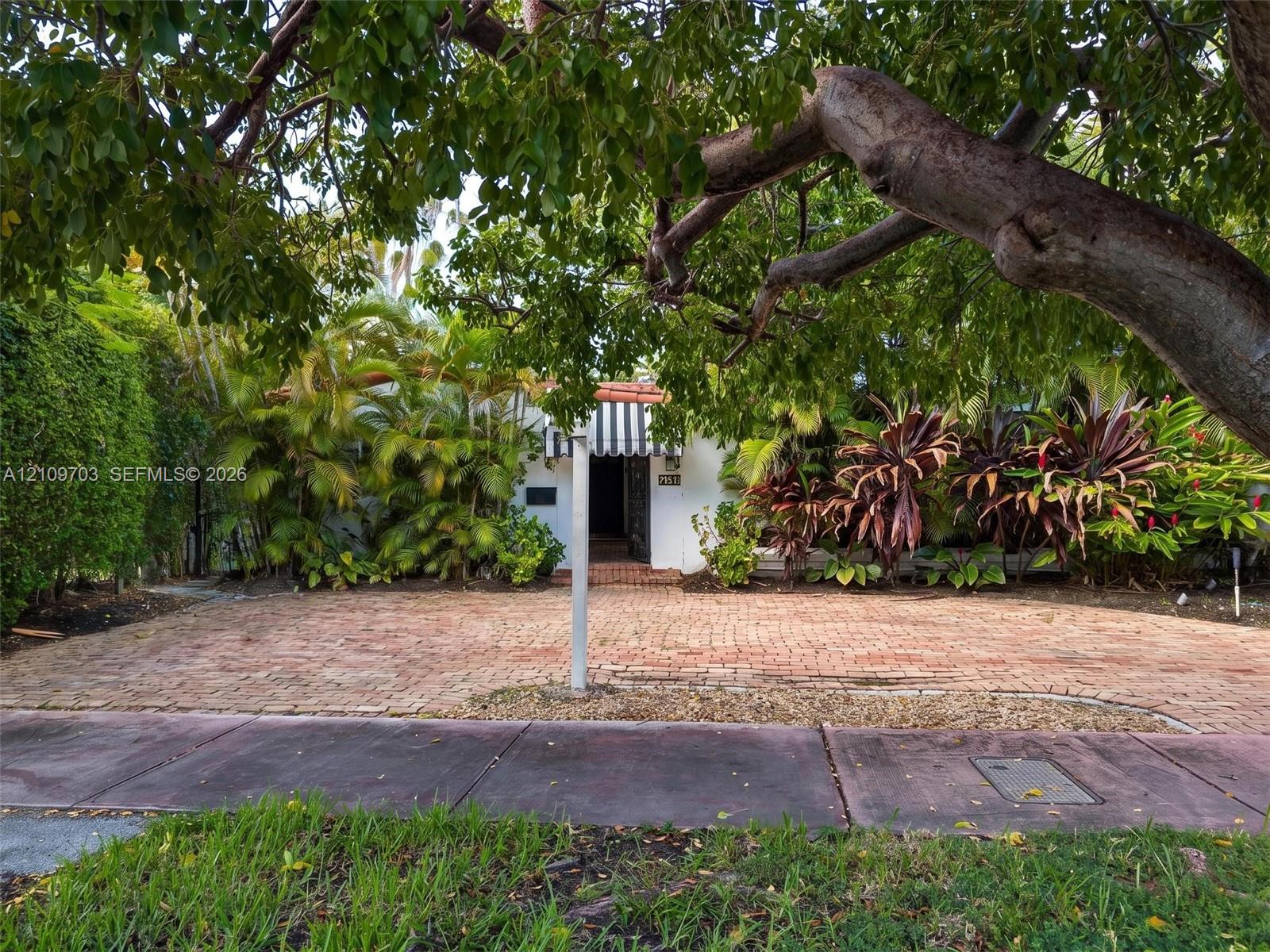 Brick-paved driveway, striped window awning, mature canopy trees, and established perimeter landscaping