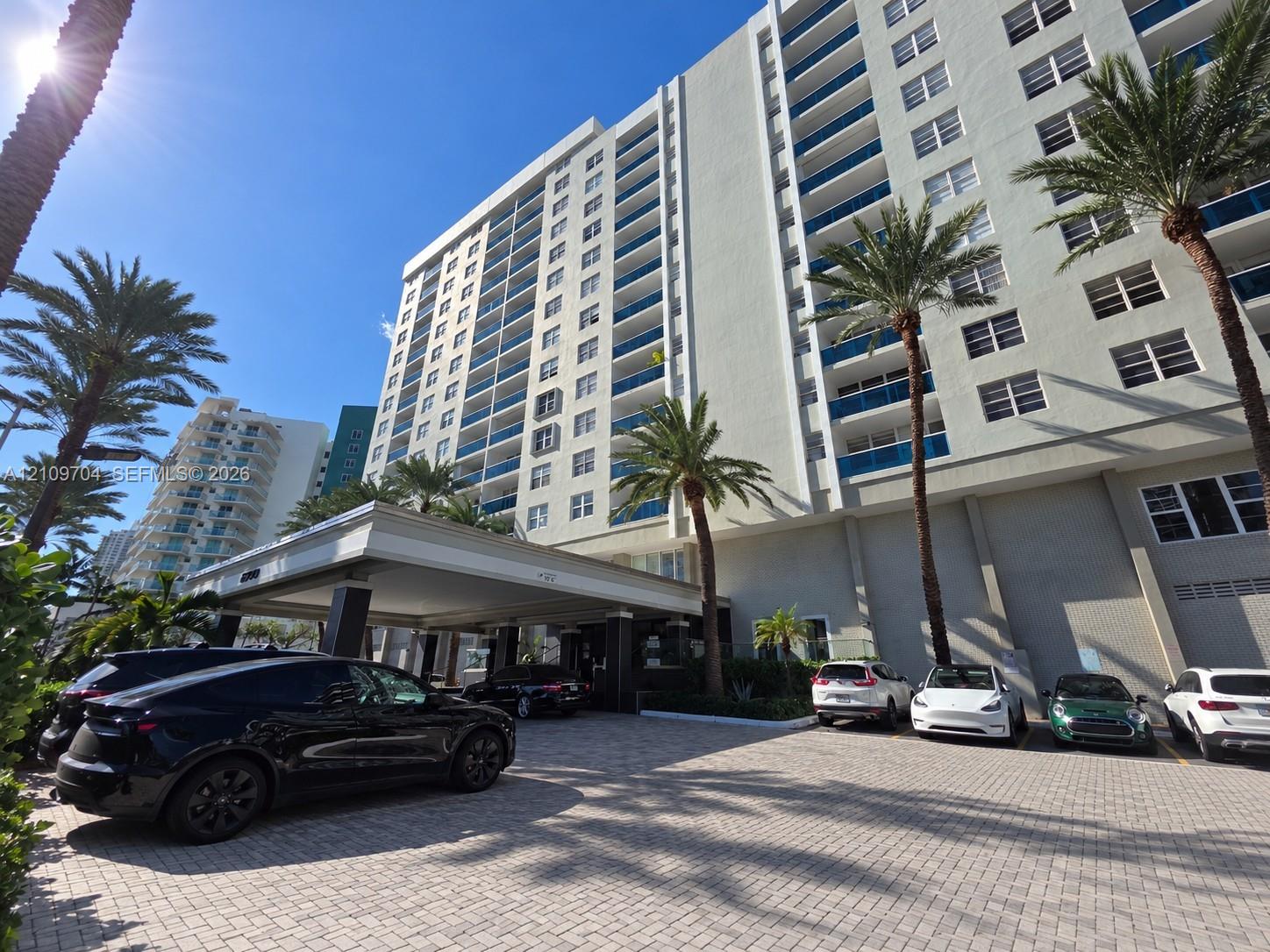 High-rise residential building featuring a paved motor court, covered porte-cochre, multi-story private balconies, and mature palm tree landscaping
