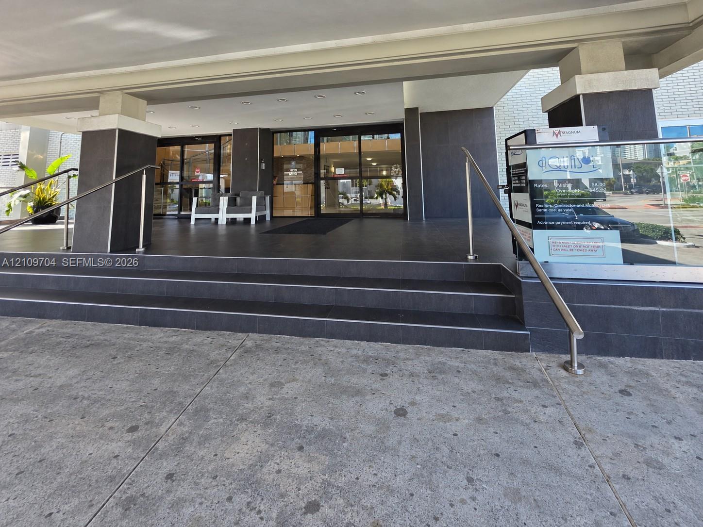 Covered building entrance featuring tiered stone steps, matte dark tile finishes, and stainless steel handrails