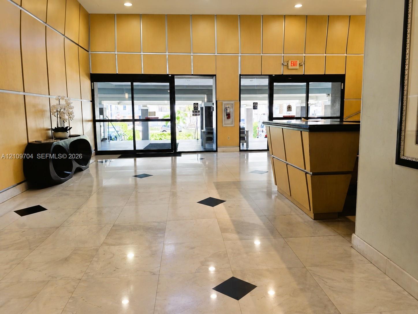 Lobby featuring polished tile flooring with inlaid stone accents, wood-paneled walls, built-in reception desk, and floor-to-ceiling glass entry doors with aluminum framing
