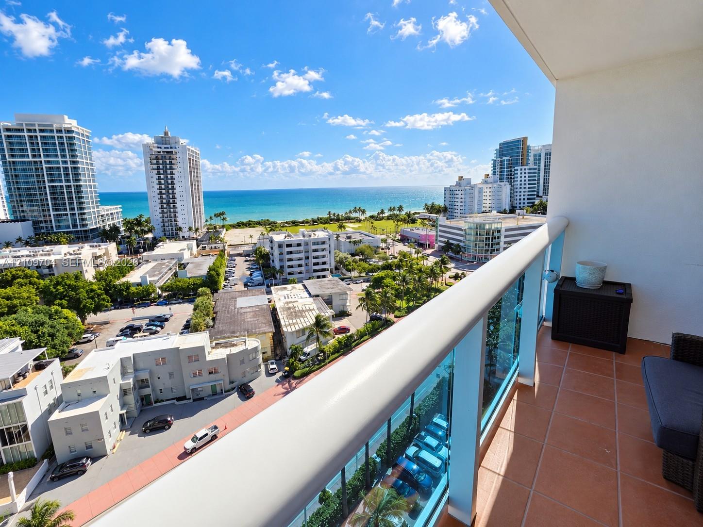 Covered balcony featuring terracotta-style tile flooring and a glass-panel railing overlooking the ocean and urban coastline