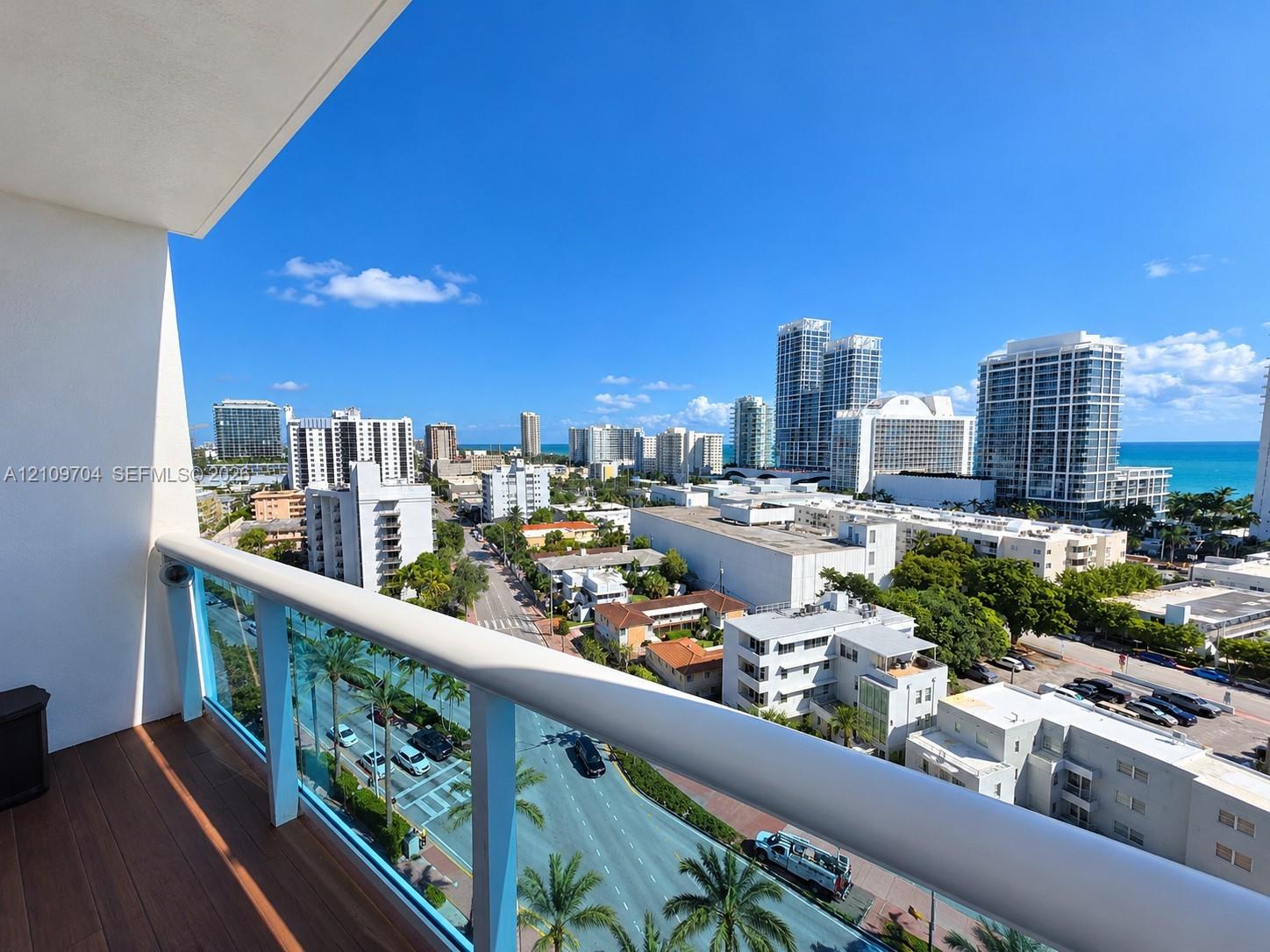 Covered balcony featuring wood-finish flooring and a glass panel railing with a metal top rail, offering an expansive cityscape and ocean horizon