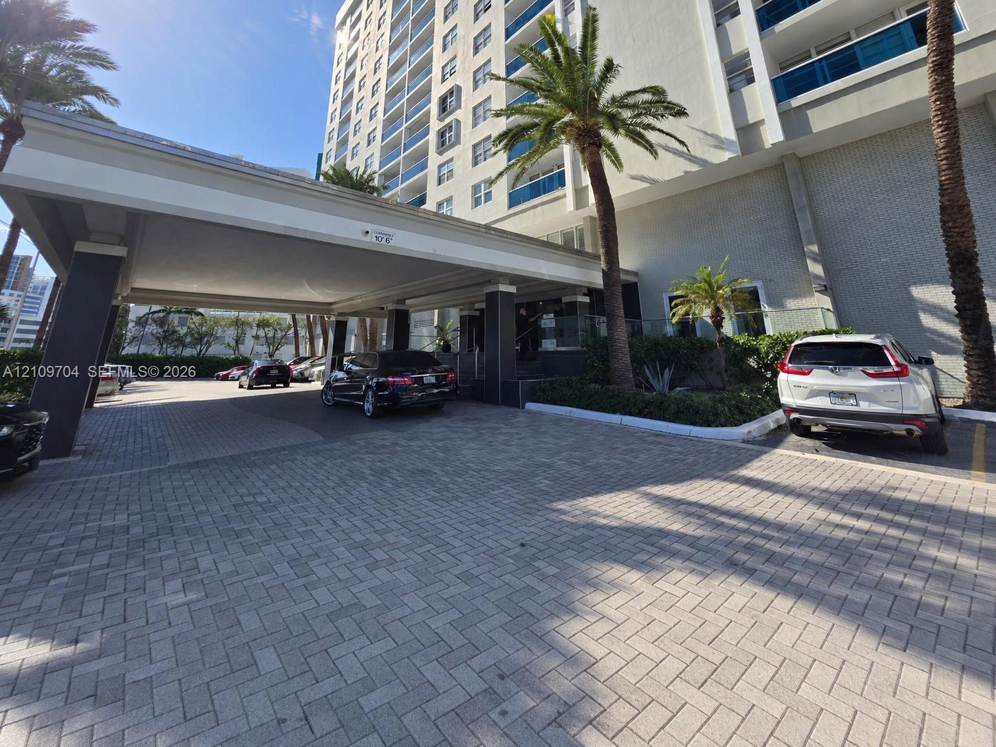 High-rise exterior featuring a paved porte-cochre, covered drop-off area, multi-story facade with glass railings, and mature palm trees