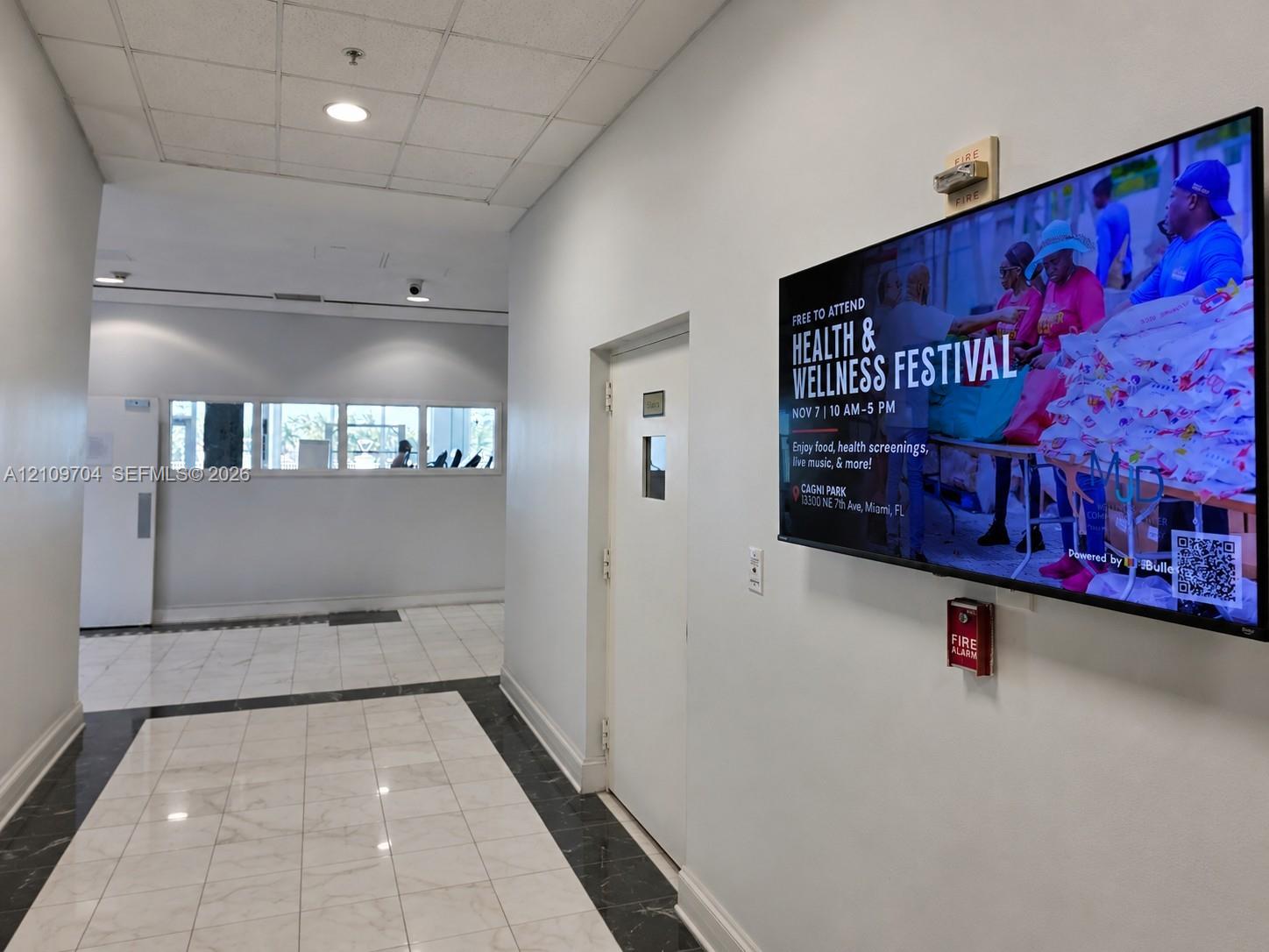 Interior corridor featuring polished stone flooring with dark inlay border, recessed ceiling lighting, and wall-mounted digital display screen