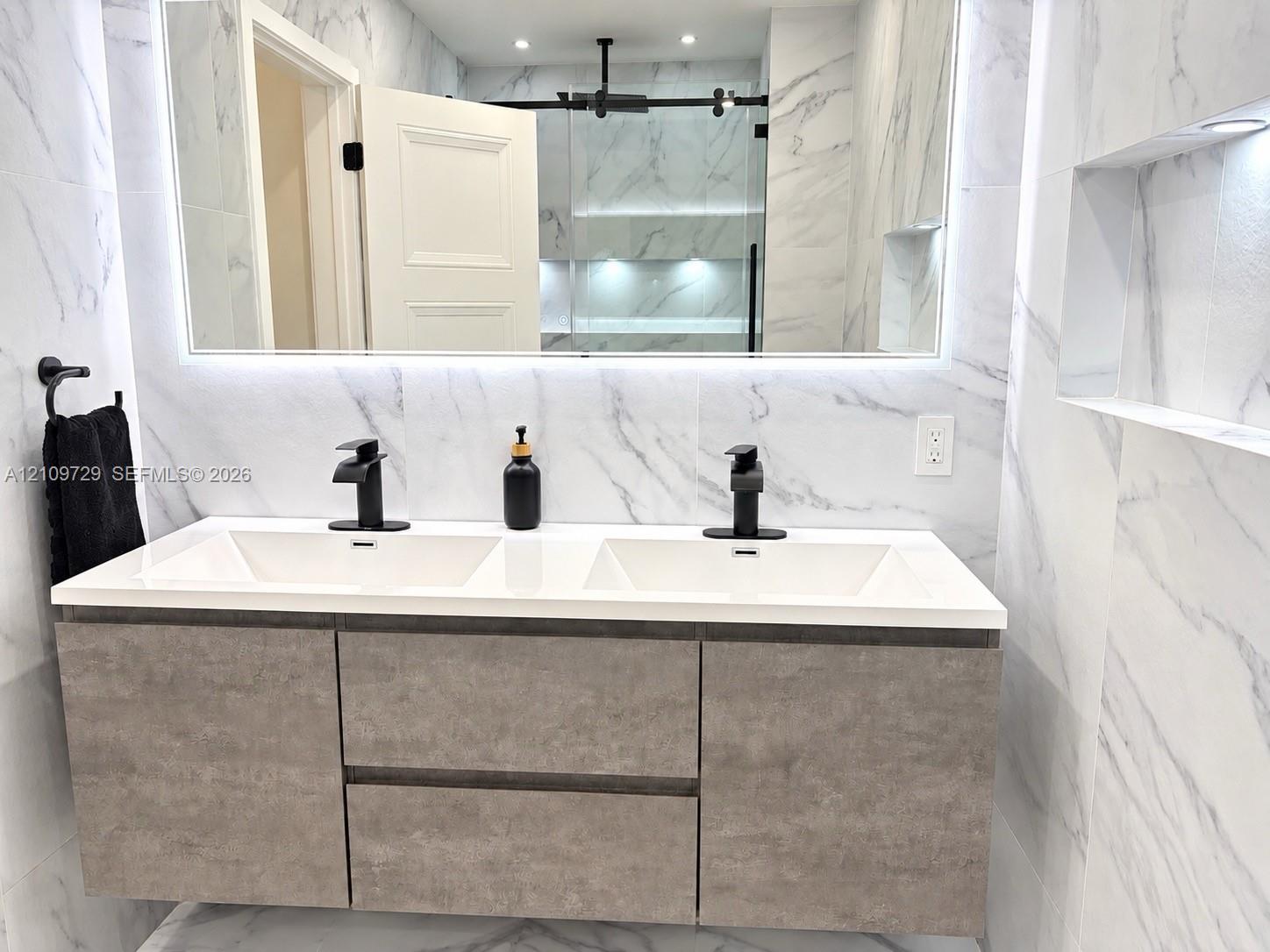 Floating double vanity with a composite countertop, dual matte black faucets, integrated LED-backlit mirror, and full-height wall tiling with recessed shelving