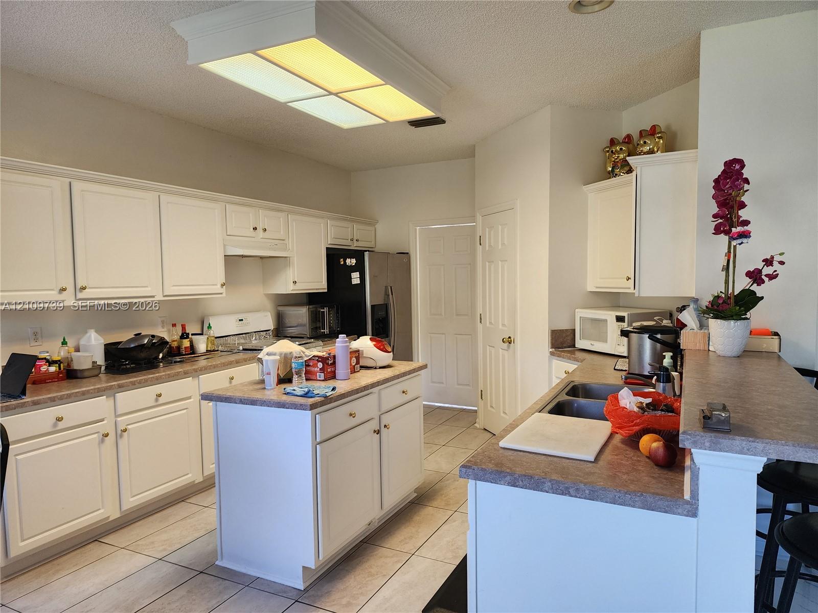 Kitchen featuring white cabinetry, central island, dual-basin stainless steel sink, solid-surface countertops, and tile flooring