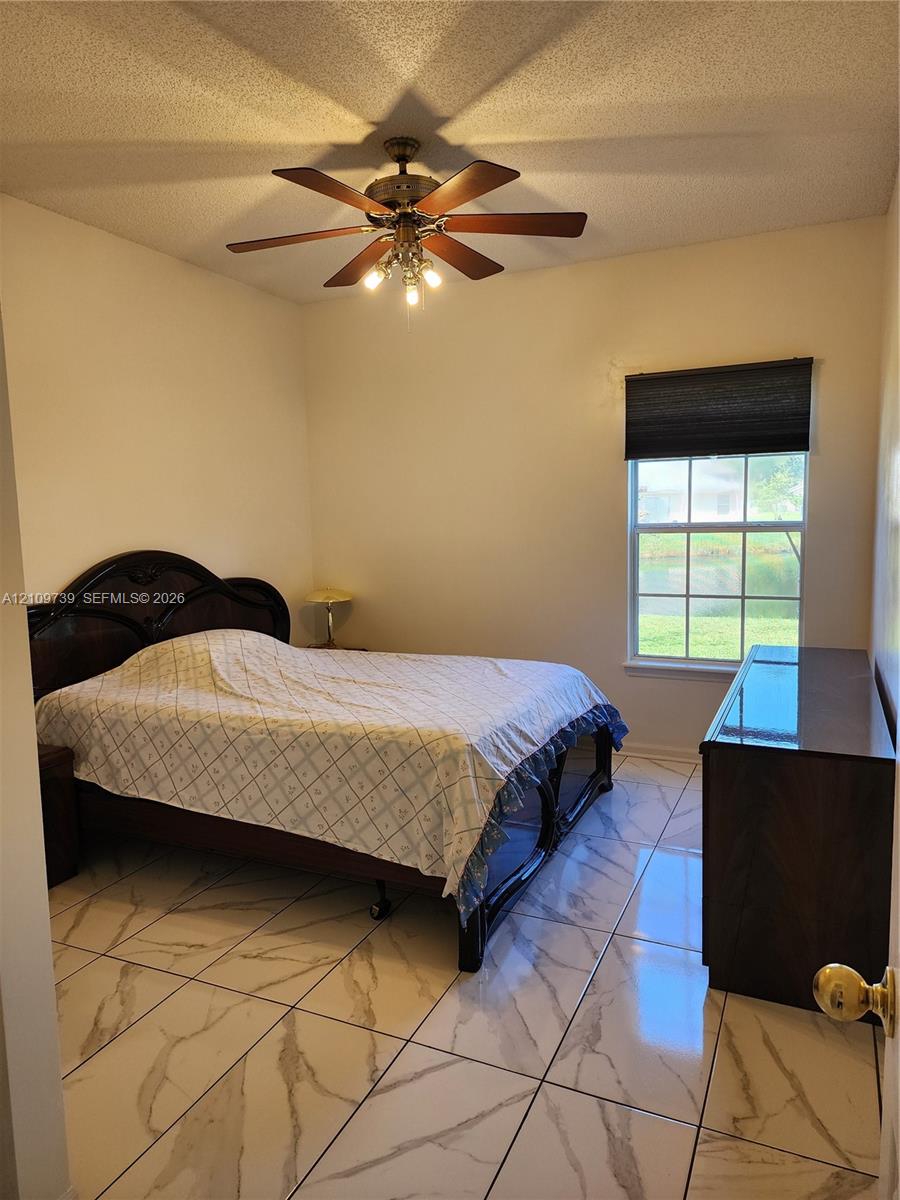 Neutral-toned room featuring large-format tile flooring with a veined pattern, multi-blade ceiling fan with integrated lighting, and a window with a retractable shade