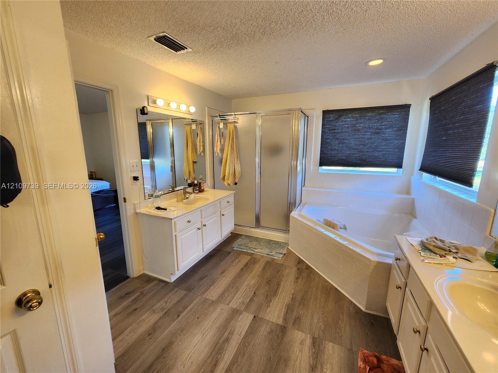 Bathroom featuring wood-finish flooring, dual white vanities, corner soaking tub with tile surround, and a glass-enclosed shower