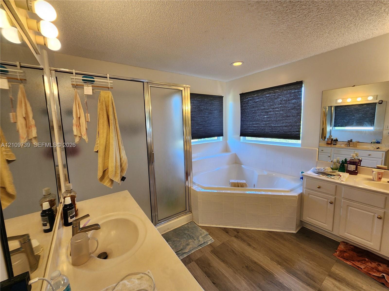 Bathroom featuring wood-finish flooring, a corner soaking tub with tiled surround, a separate glass-enclosed shower, dual vanity cabinets, and a framed mirror