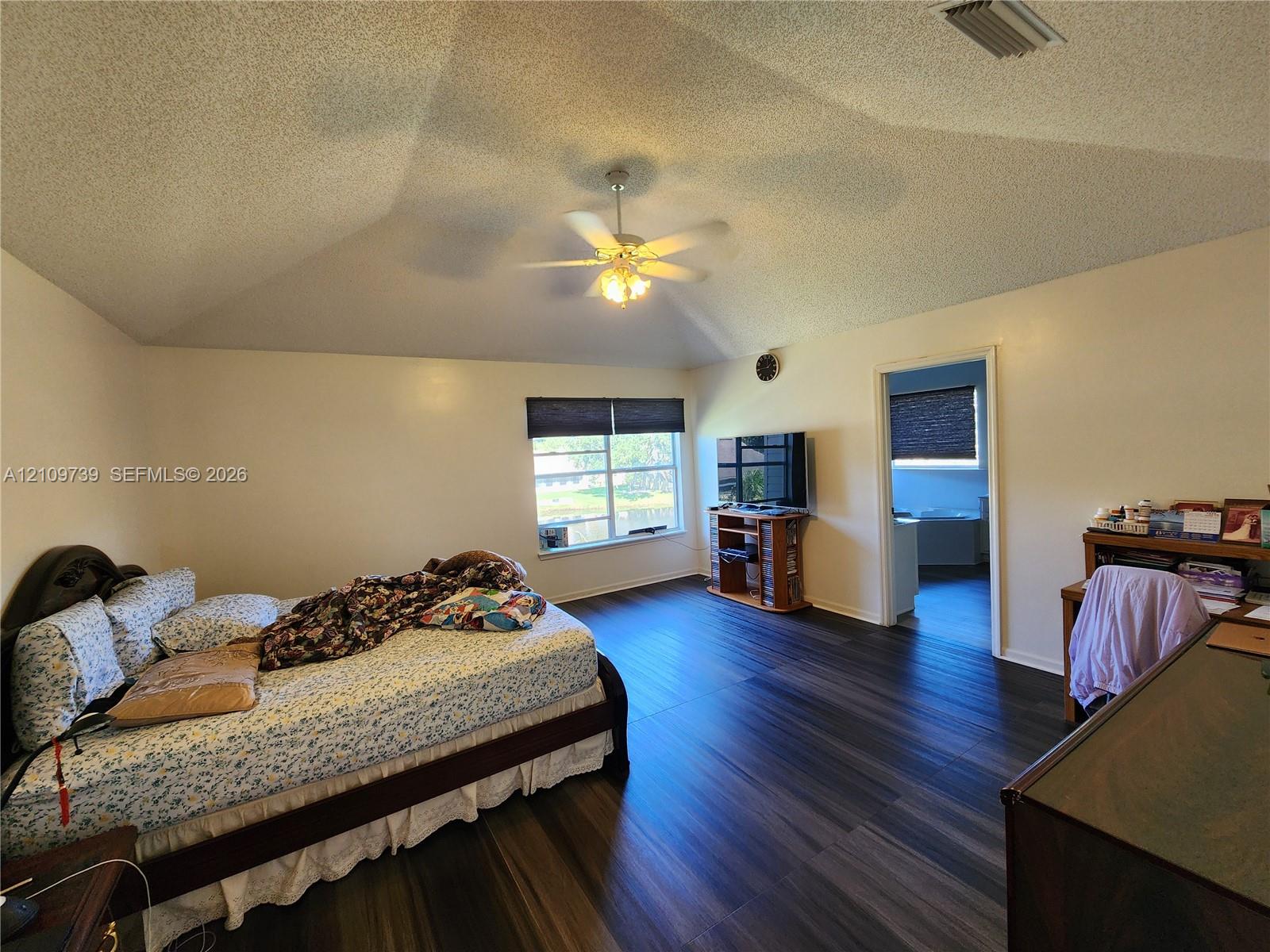 Vaulted ceiling with a centrally mounted ceiling fan, dark wood-finish flooring, and large window with roller shades