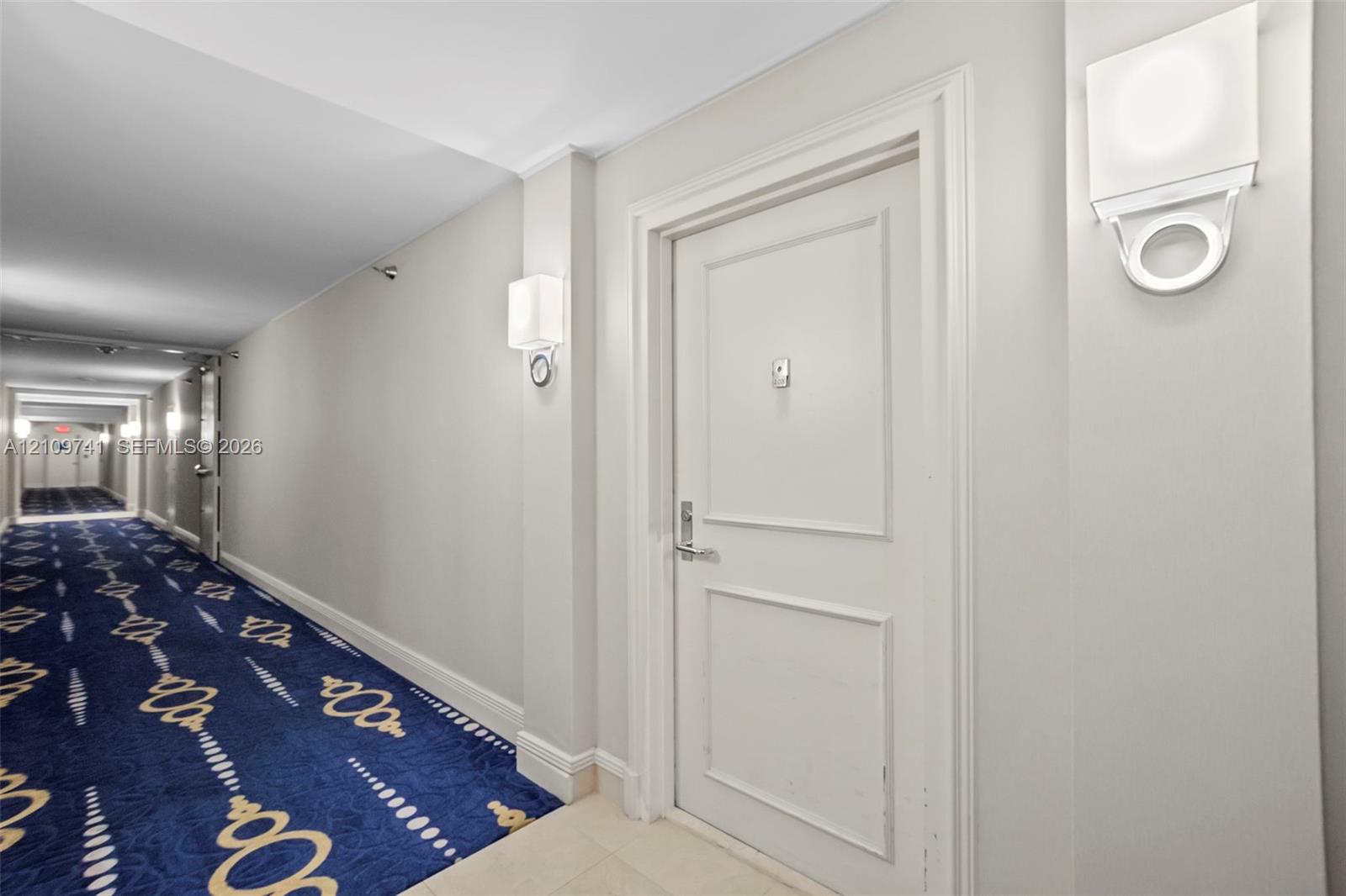 Hallway featuring patterned carpet flooring, white paneled interior doors, and modern wall-mounted light fixtures