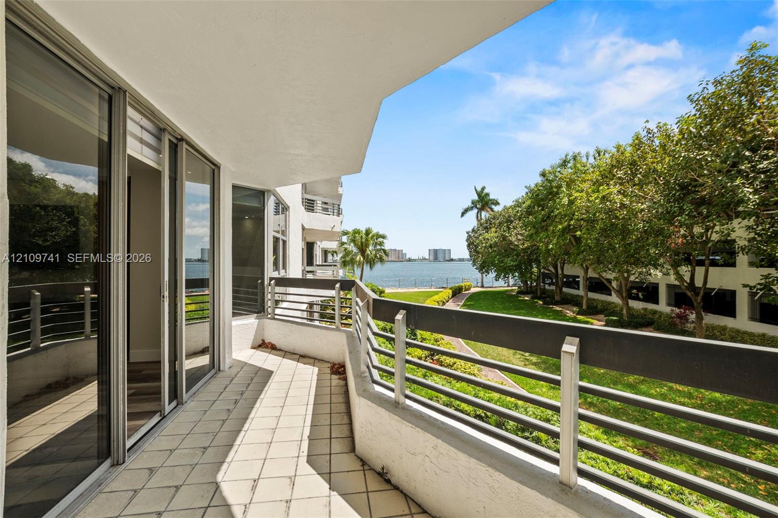 Covered balcony featuring tile flooring, metal railings, and sliding glass door entry, with water frontage and mature landscaping