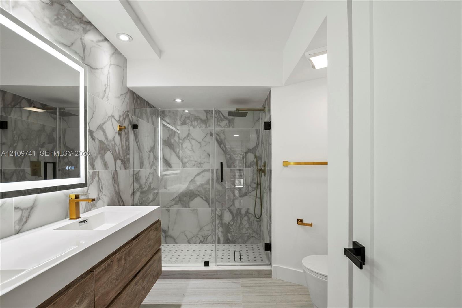 Floating wood-finish vanity with an integrated sink, backlit LED mirror, walk-in shower with stone-patterned tile, and gold-tone fixtures