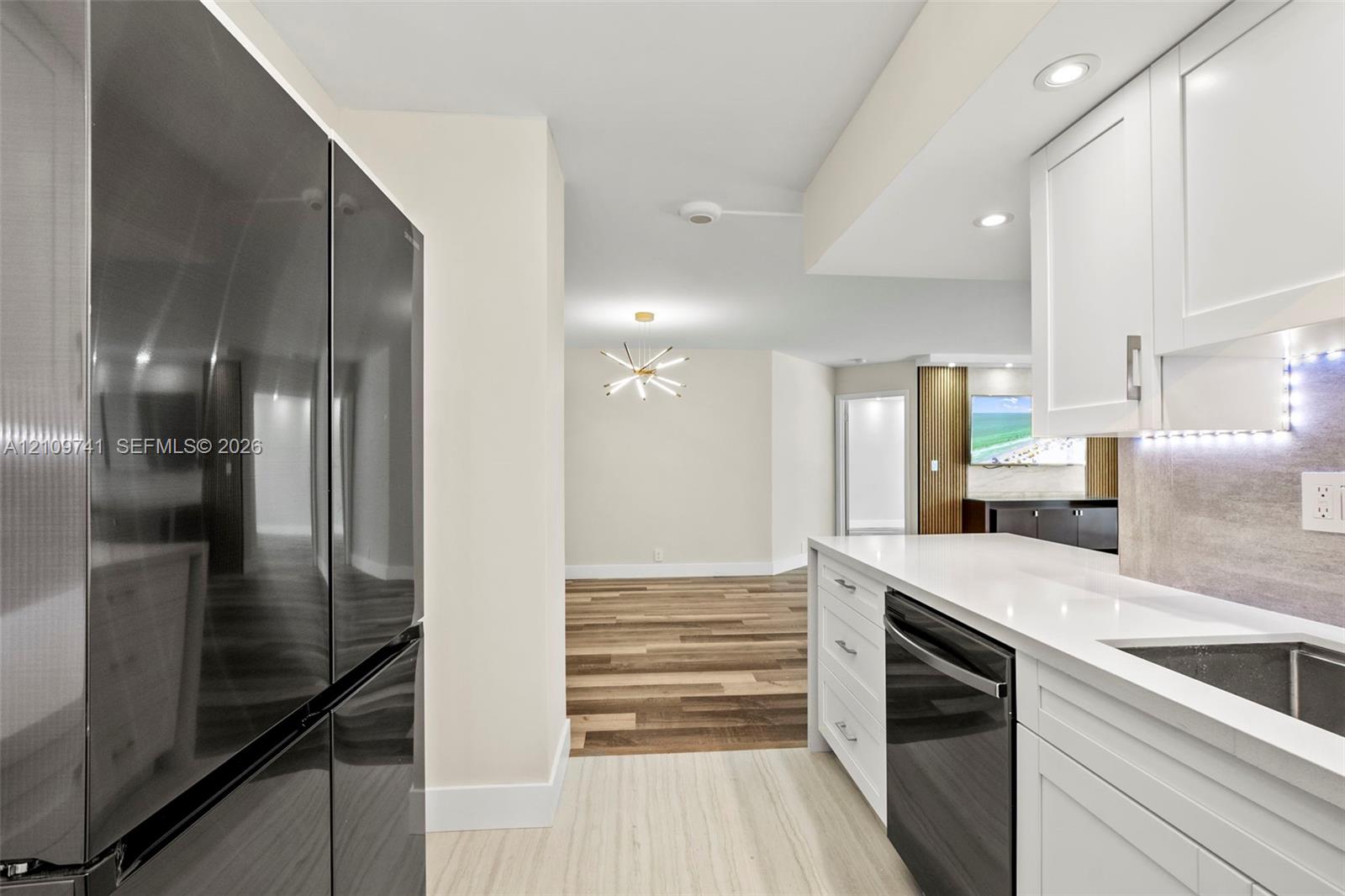 Kitchen featuring white shaker-style cabinetry, quartz countertops, integrated under-cabinet lighting, a stainless steel dishwasher, and a full-size dark-finish refrigerator Kitchen featuring white shaker-style cabinetry, quartz countertops, integrated under-cabinet lighting, a stainless steel dishwasher, and a full-size dark-finish refrigerator