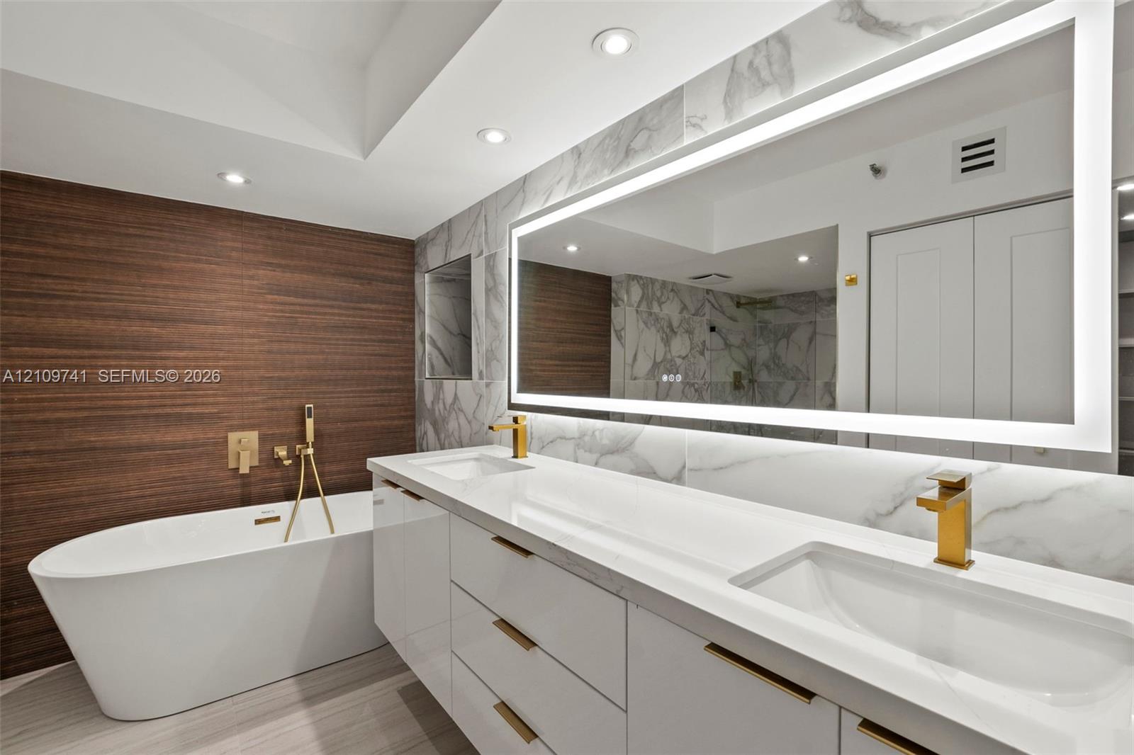 Freestanding soaking tub with wall-mounted gold-toned hardware, white vanity with integrated double sinks, large LED-backlit mirror, wood-finish feature wall, and stone-finish wall tile