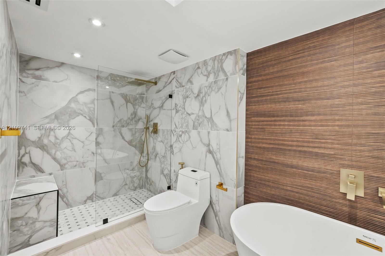 Full-height stone-patterned tile shower enclosure with integrated bench and frameless glass door, freestanding soaking tub, wood-finish textured accent wall, and gold-toned wall-mounted plumbing fixtures