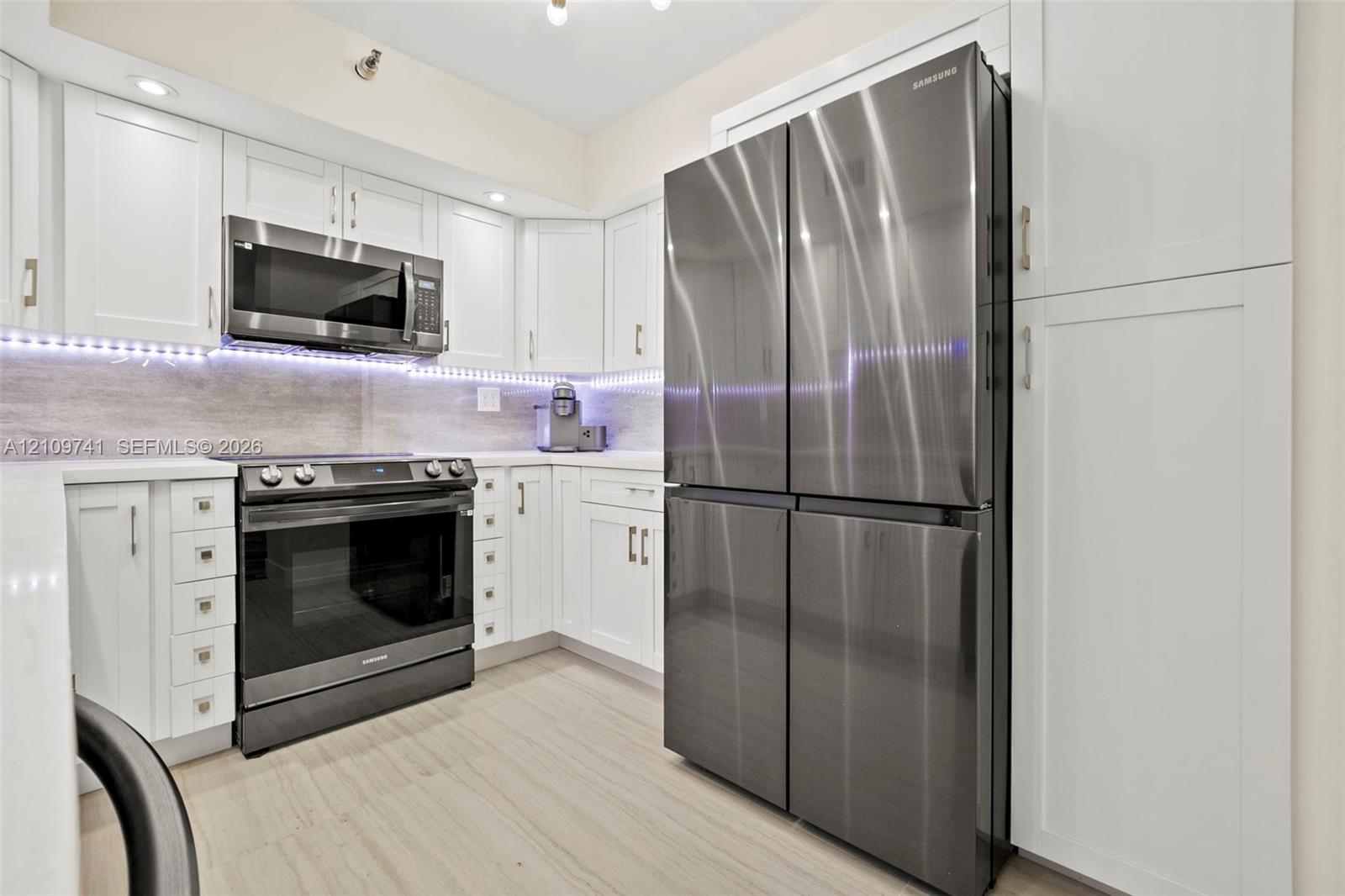 Kitchen featuring white shaker-style cabinetry, stainless steel appliances, stone-surface countertops, under-cabinet lighting, and wood-finish flooring Kitchen featuring white shaker-style cabinetry, stainless steel appliances, stone-surface countertops, under-cabinet lighting, and wood-finish flooring