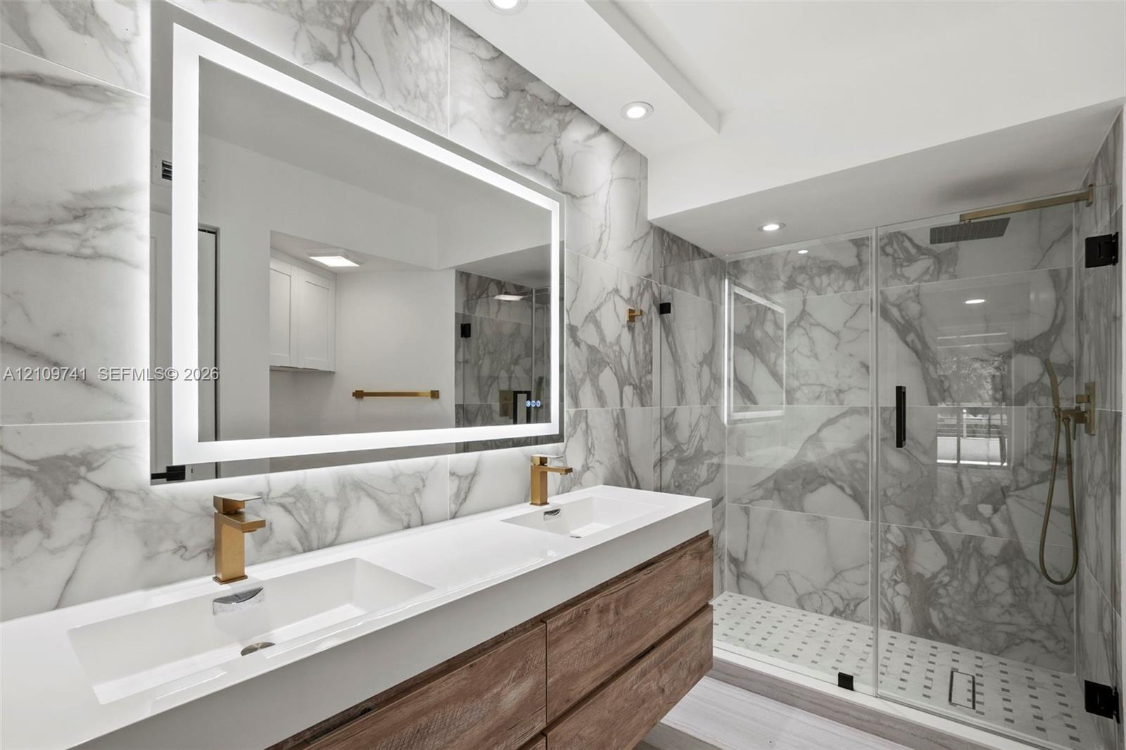 Floating dual-sink vanity with wood-finish cabinetry, backlit LED mirror, large-format stone-patterned tile wall, and glass-enclosed shower with gold-tone hardware