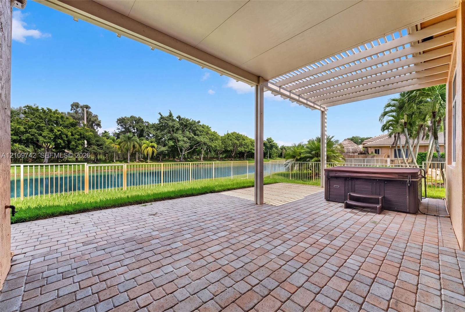 Covered paver patio featuring an attached pergola, built-in spa, and metal perimeter fencing overlooking the water