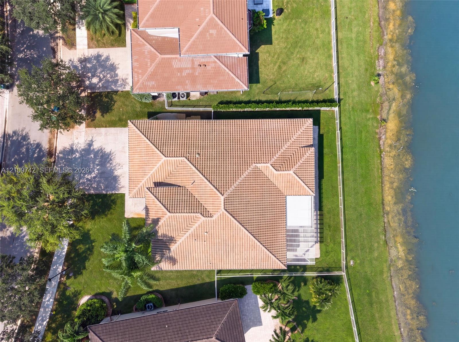 Aerial perspective of a residential property featuring a multi-hipped tile roof, concrete driveway, and expansive lawn area with perimeter fencing along the waterfront