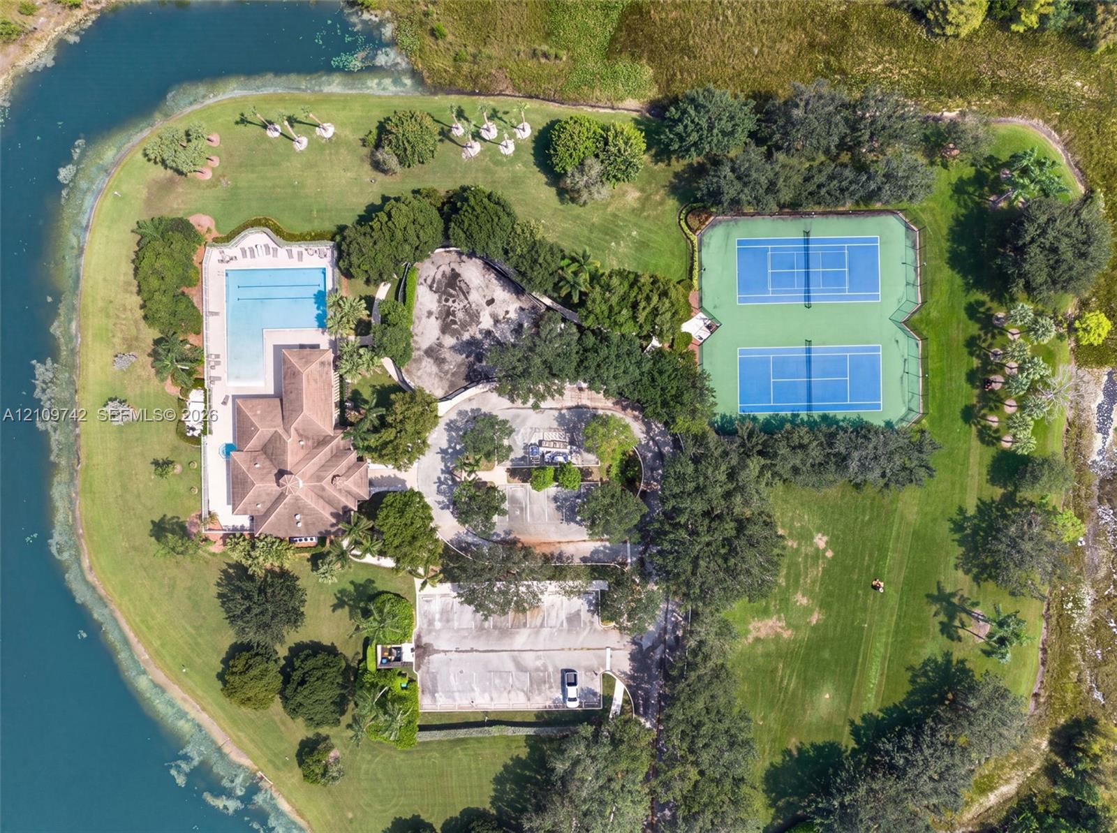 Aerial perspective of a multi-acre property featuring a swimming pool, two hard-court tennis courts, expansive manicured lawn, and water frontage