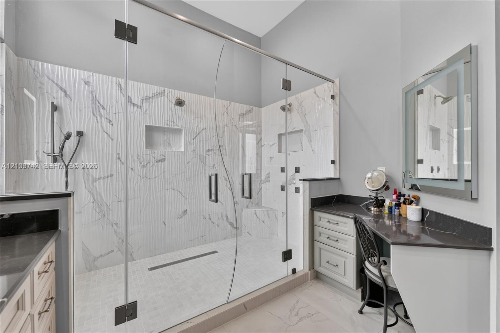 Walk-in frameless glass shower featuring textured tile surround, built-in bench, dual shower heads, and multiple body sprays, complemented by dual vanities with dark stone countertops and an illuminated mirror