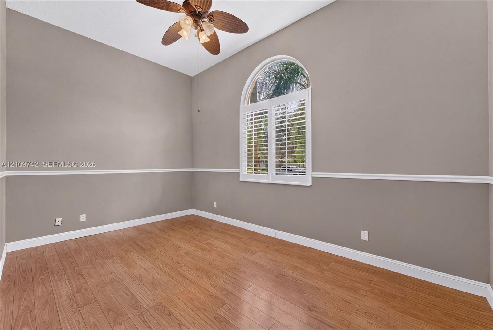 Room featuring wood-finish flooring, an arched window with plantation shutters, decorative chair rail molding, and a multi-blade ceiling fan