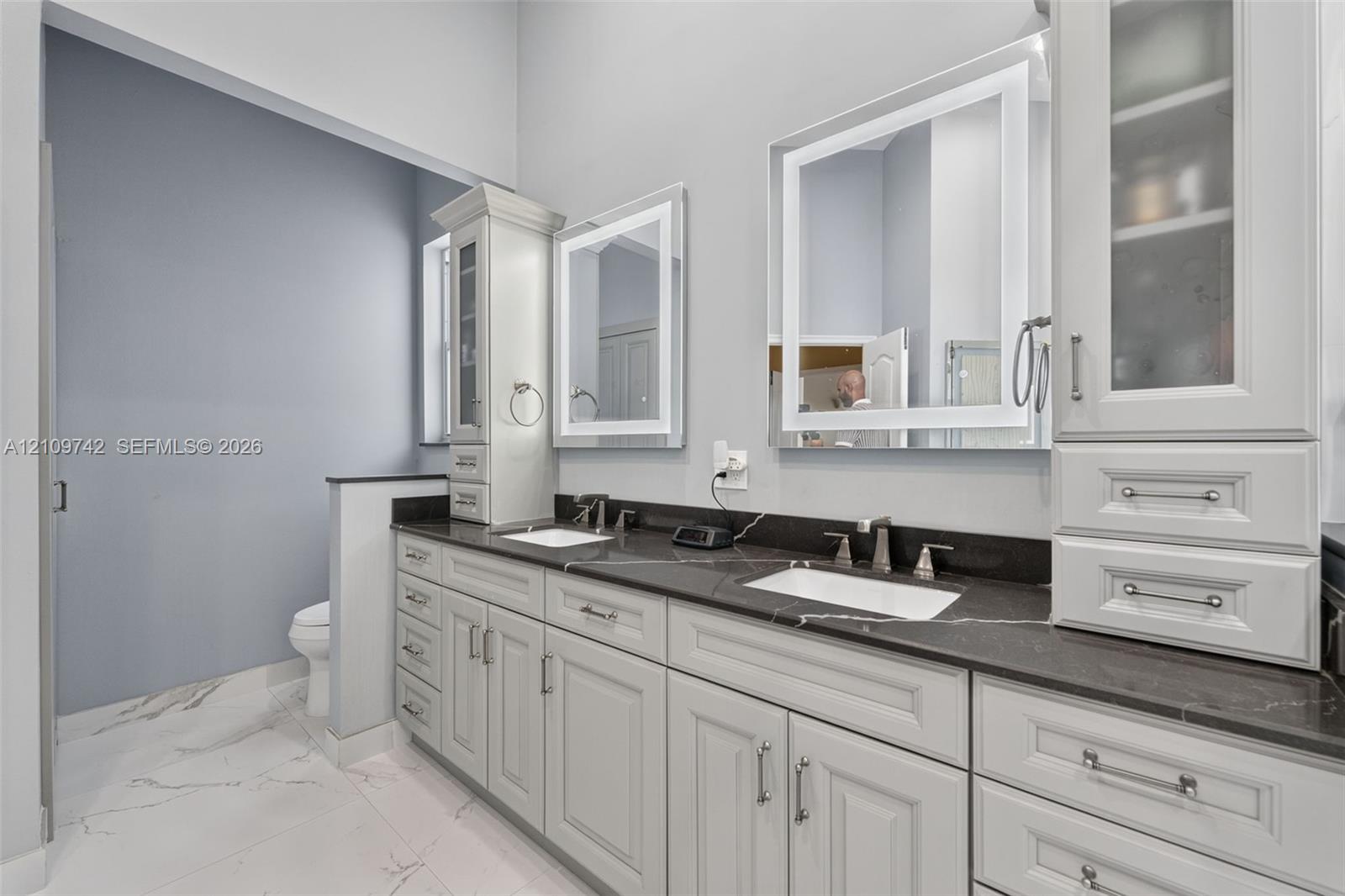 Dual-sink vanity with dark stone countertop, white shaker-style cabinetry, illuminated wall mirrors, and porcelain tile flooring