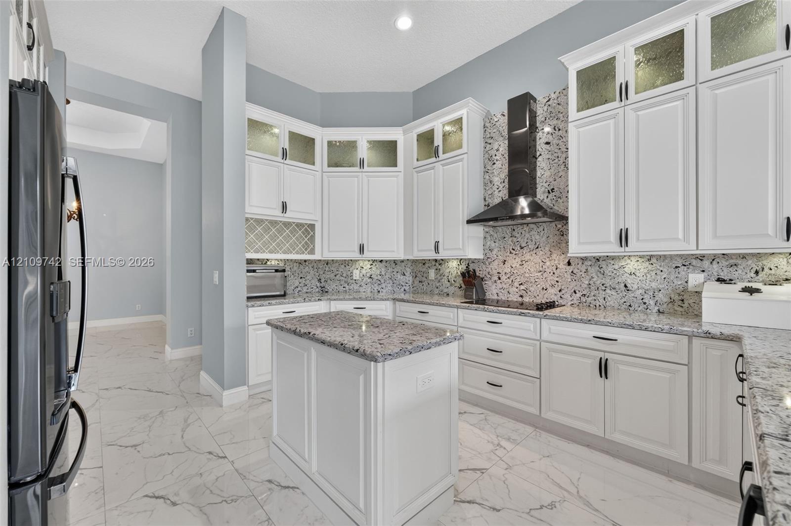 Kitchen featuring white shaker-style cabinetry with glass-panel uppers, patterned stone backsplash, center island, integrated cooktop with stainless steel range hood, and tile flooring