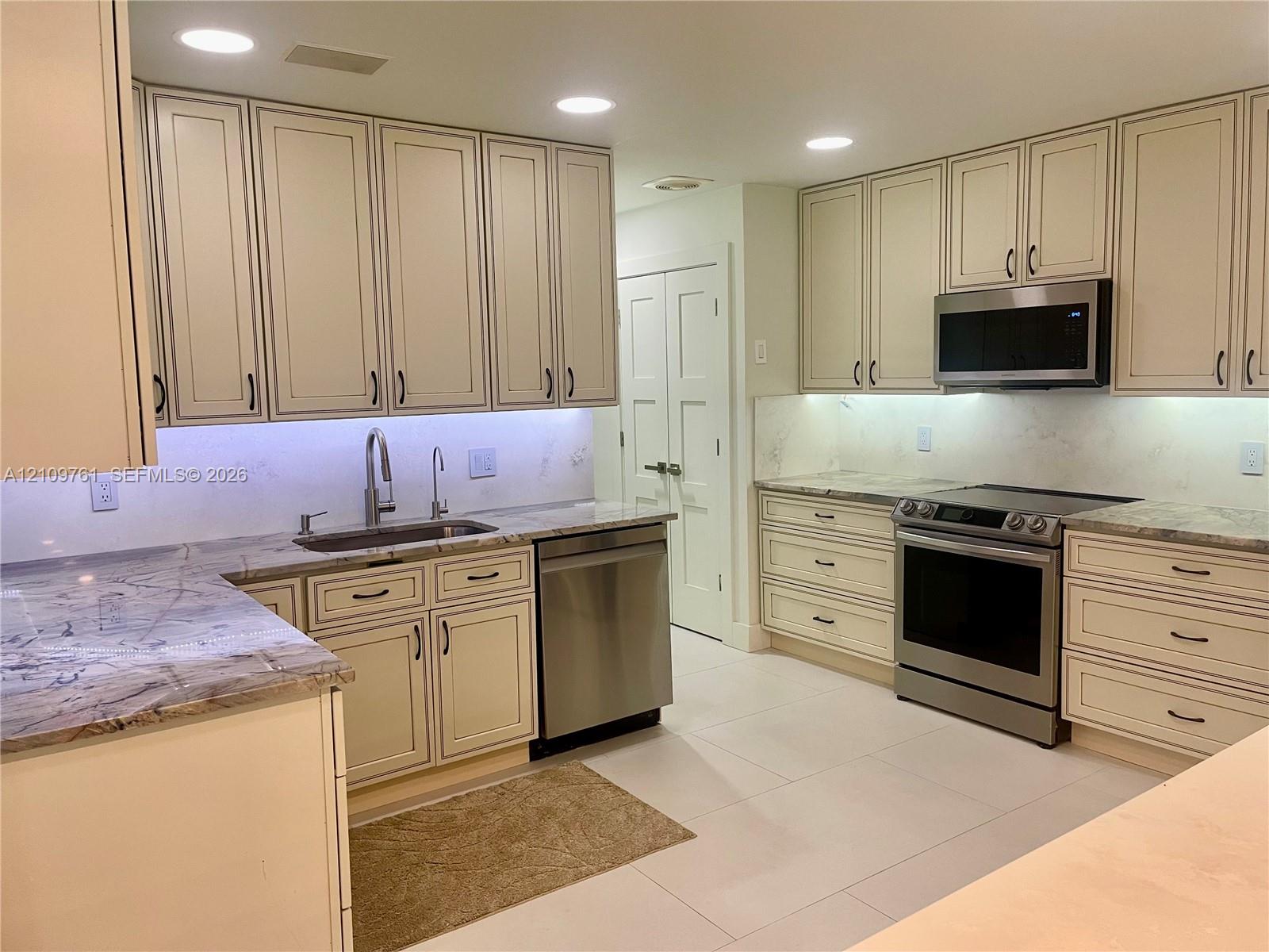 Kitchen featuring off-white cabinetry, stone-finish countertops, stainless steel appliances, under-cabinet lighting, and large-format tile flooring Kitchen featuring off-white cabinetry, stone-finish countertops, stainless steel appliances, under-cabinet lighting, and large-format tile flooring