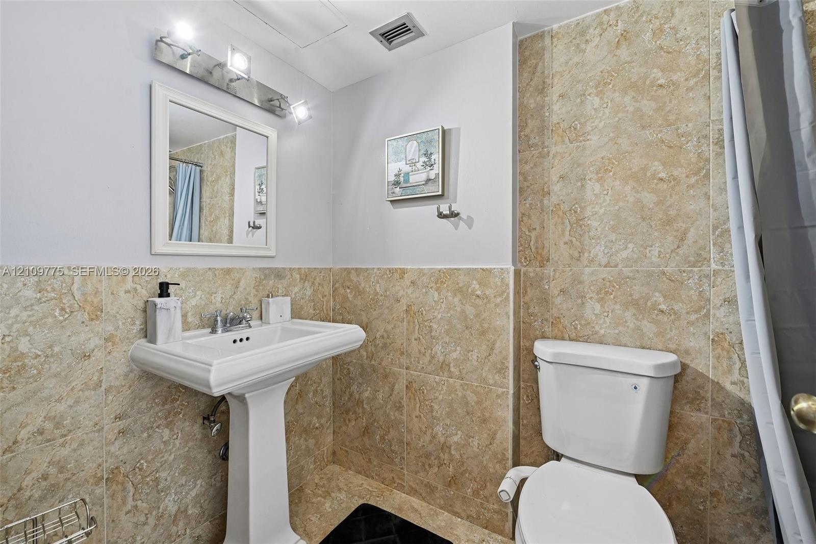 Pedestal sink with a widespread faucet, framed wall-mounted mirror, three-light vanity fixture, and full-height stone-finish tile wall surfacing
