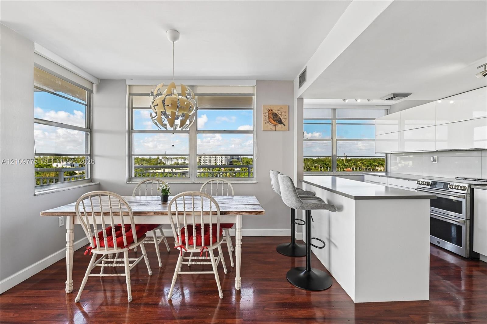 Kitchen featuring high-gloss white cabinetry, stainless steel appliances, stone-surface breakfast bar, and wood-finish flooring with large-scale windows