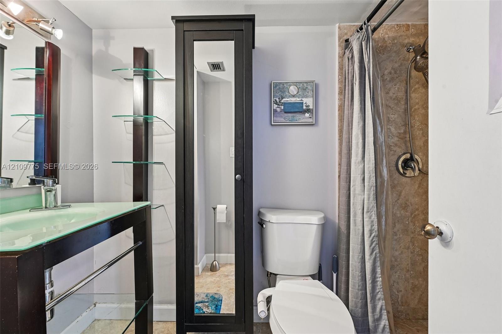 Bathroom featuring a glass-topped vanity with an integrated basin, a tall dark-finish built-in storage cabinet with a mirrored door, glass shelving, and a tiled shower stall with a hand-held shower head