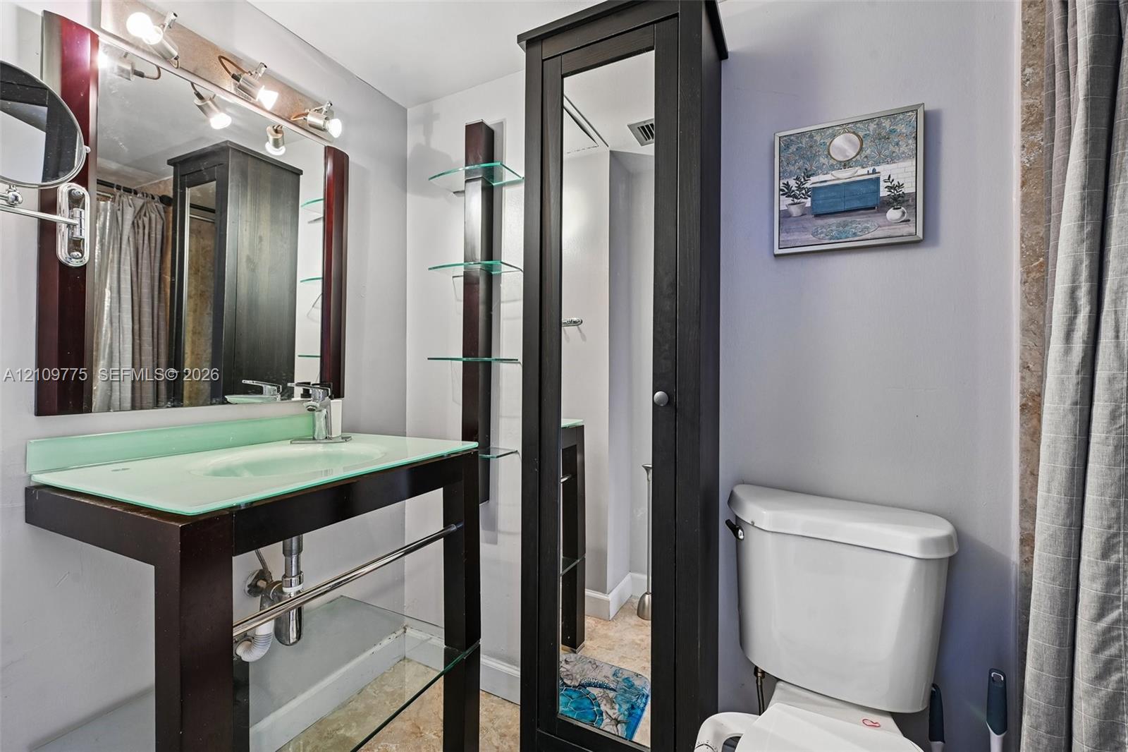 Bathroom featuring a floating vanity with a glass countertop, integrated sink, polished metal hardware, a tall mirrored storage cabinet, and built-in glass shelving