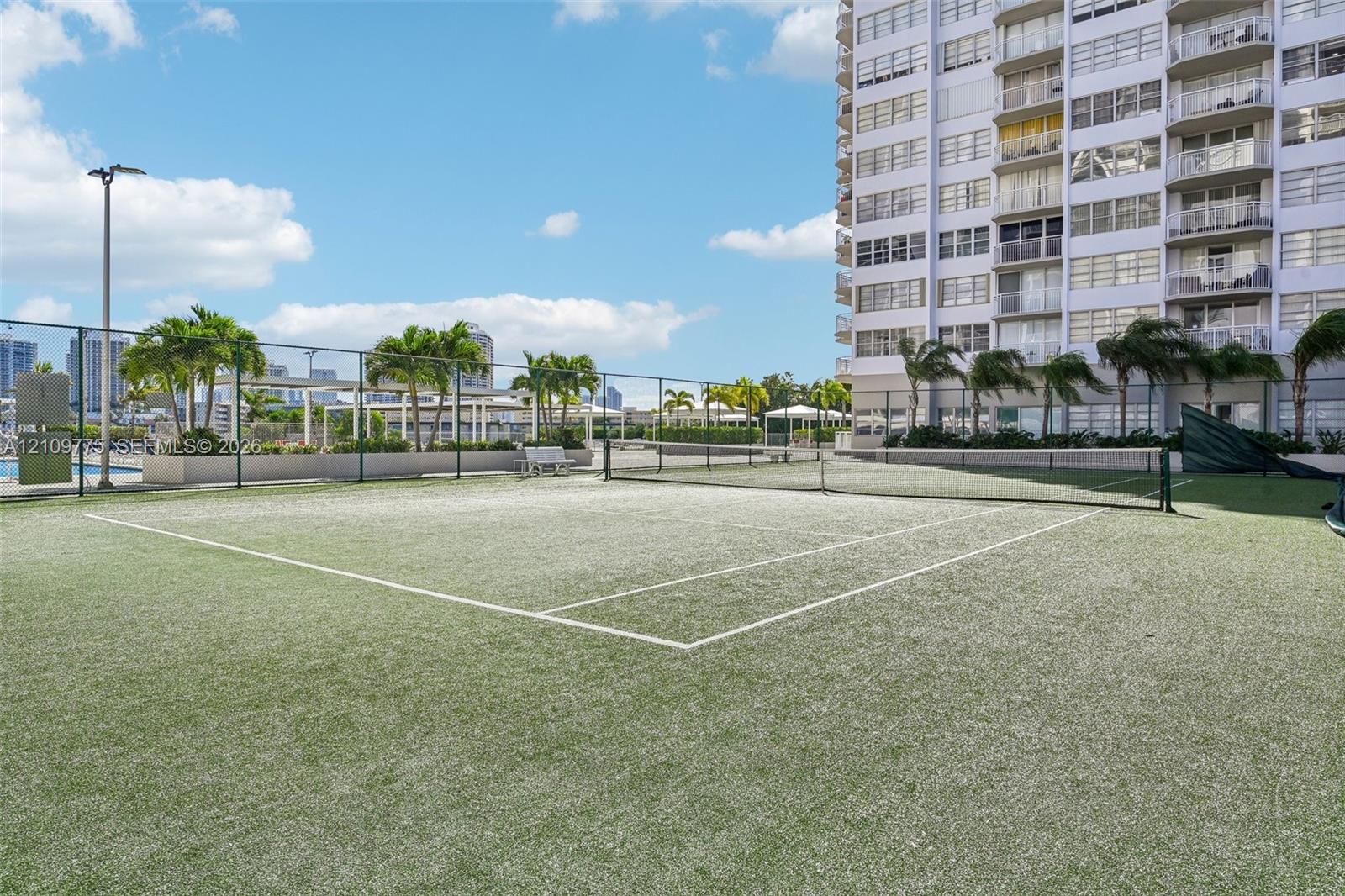 Tennis court with synthetic turf surface, enclosed by chain-link fencing, featuring overhead lighting and perimeter landscaping with palm trees