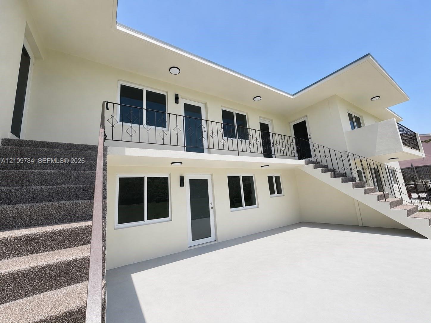 Two-story exterior featuring light-toned stucco, an upper-level balcony with decorative metal railing, concrete stairs, and multiple entry points with glass-paneled doors Two-story exterior featuring light-toned stucco, an upper-level balcony with decorative metal railing, concrete stairs, and multiple entry points with glass-paneled doors