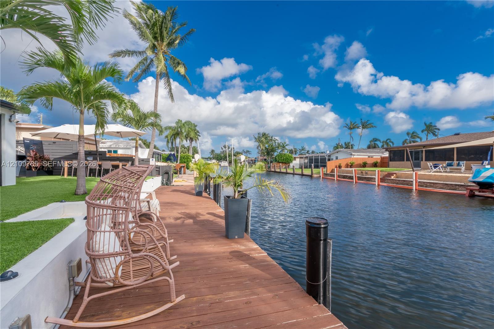 Waterfront property featuring a wooden deck, seawall, artificial turf area, and mature palm trees