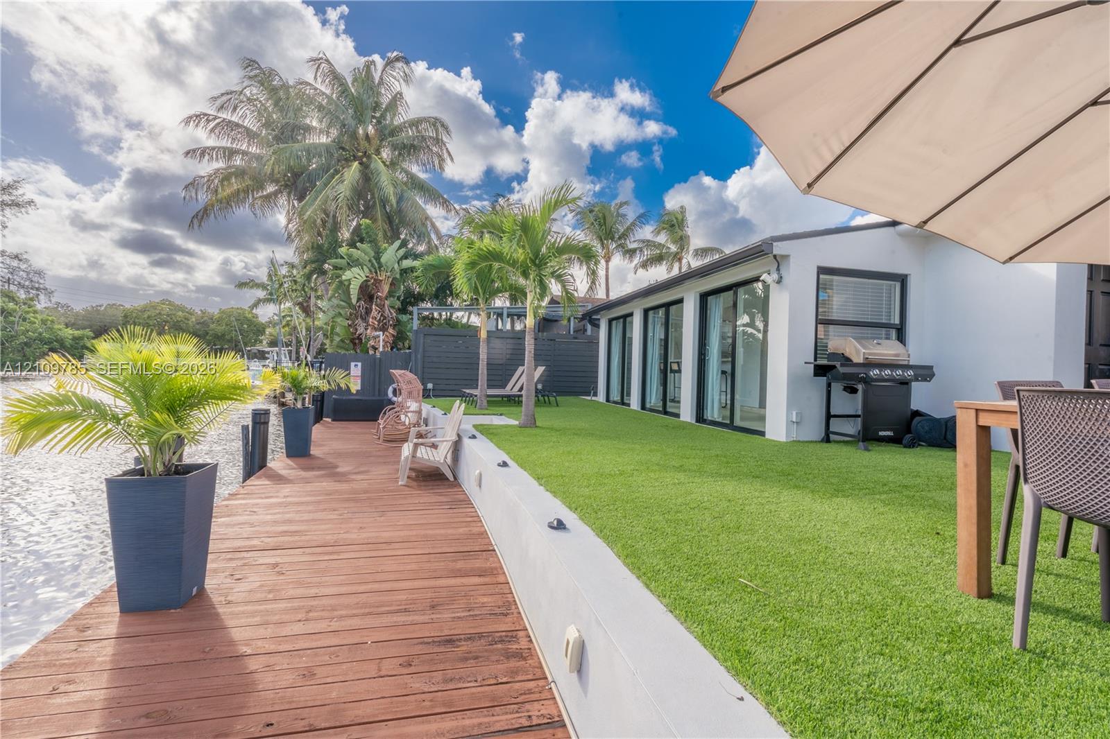 Waterfront backyard featuring a deck, synthetic turf lawn, concrete retaining wall, and sliding impact glass doors