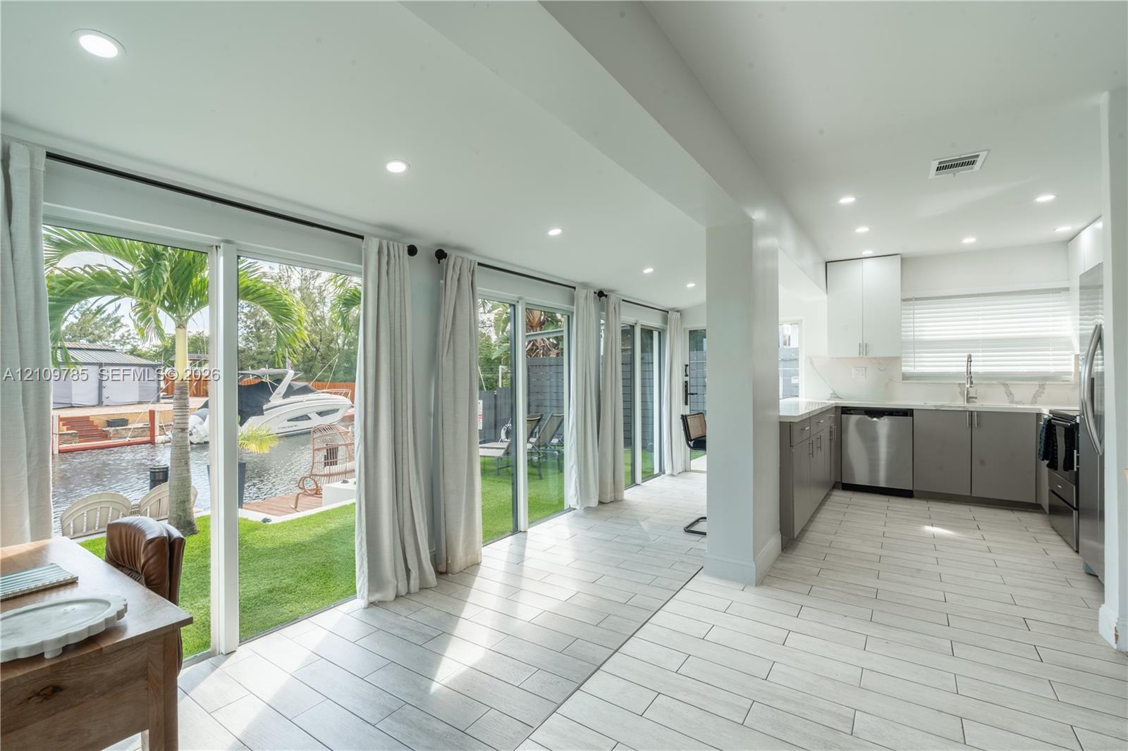Kitchen featuring stainless steel appliances, white upper cabinetry, grey lower cabinetry, and recessed lighting, paired with floor-to-ceiling glass sliding doors opening to a waterfront lawn and boat dock