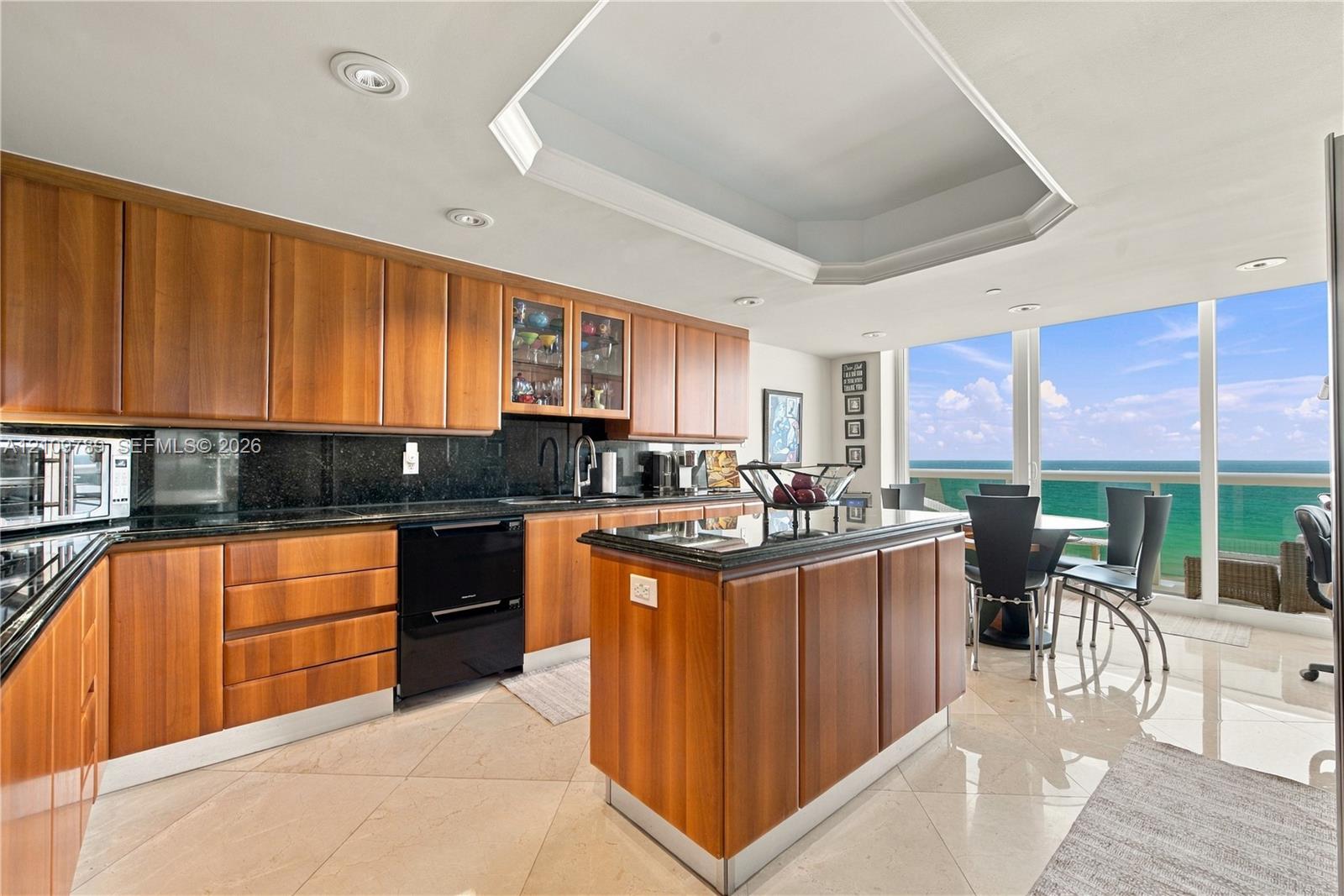 Kitchen featuring custom wood cabinetry, dark stone countertops, central island, recessed tray ceiling, and floor-to-ceiling windows with ocean scenery