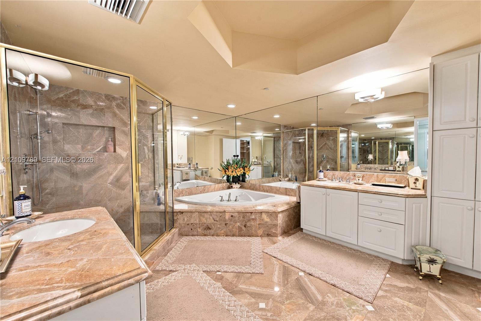 Bathroom featuring stone-surface vanity countertops, an oversized corner soaking tub with a stone surround, a glass-enclosed shower, expansive wall-to-wall mirror, and floor-to-ceiling built-in cabinetry
