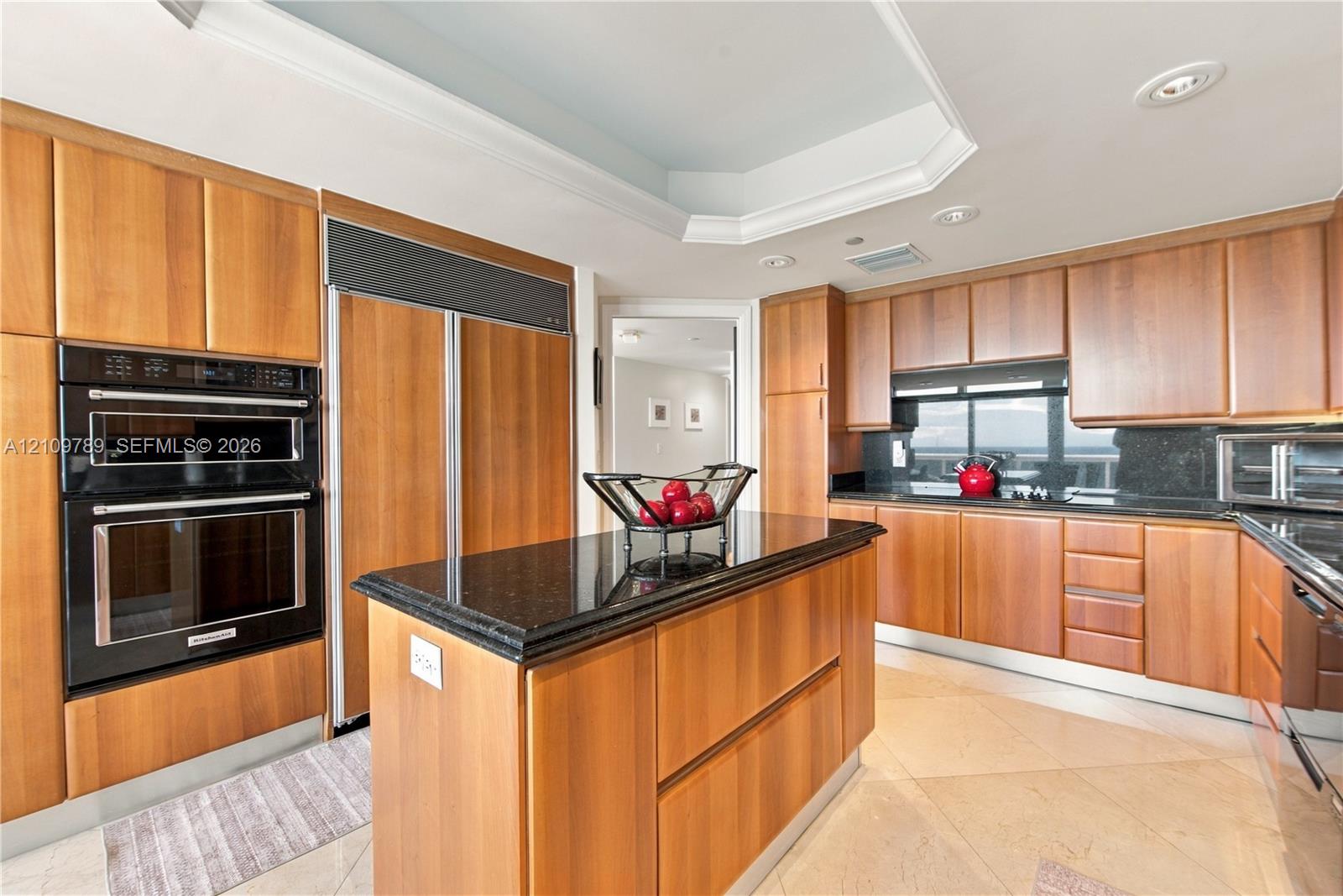 Kitchen featuring wood cabinetry, stone countertops, central island, built-in double oven, and recessed tray ceiling
