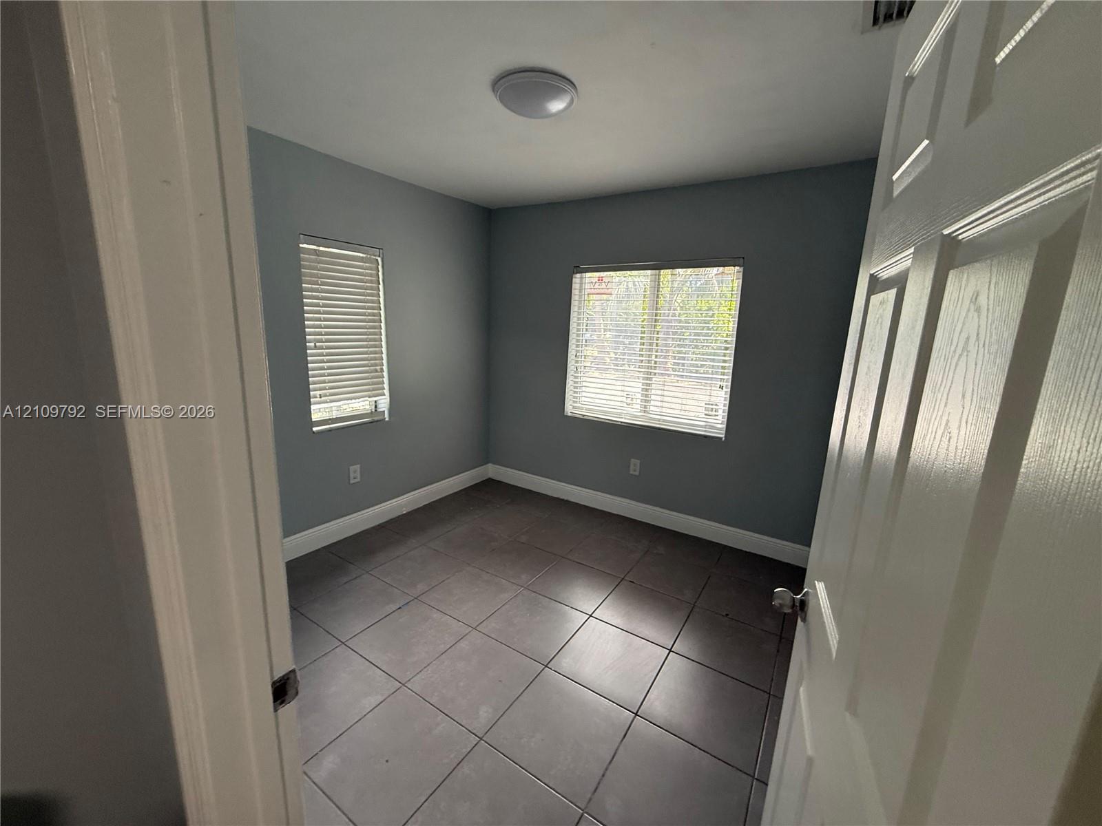 Room featuring large-format ceramic tile flooring, neutral grey wall paint, and two windows equipped with horizontal blinds