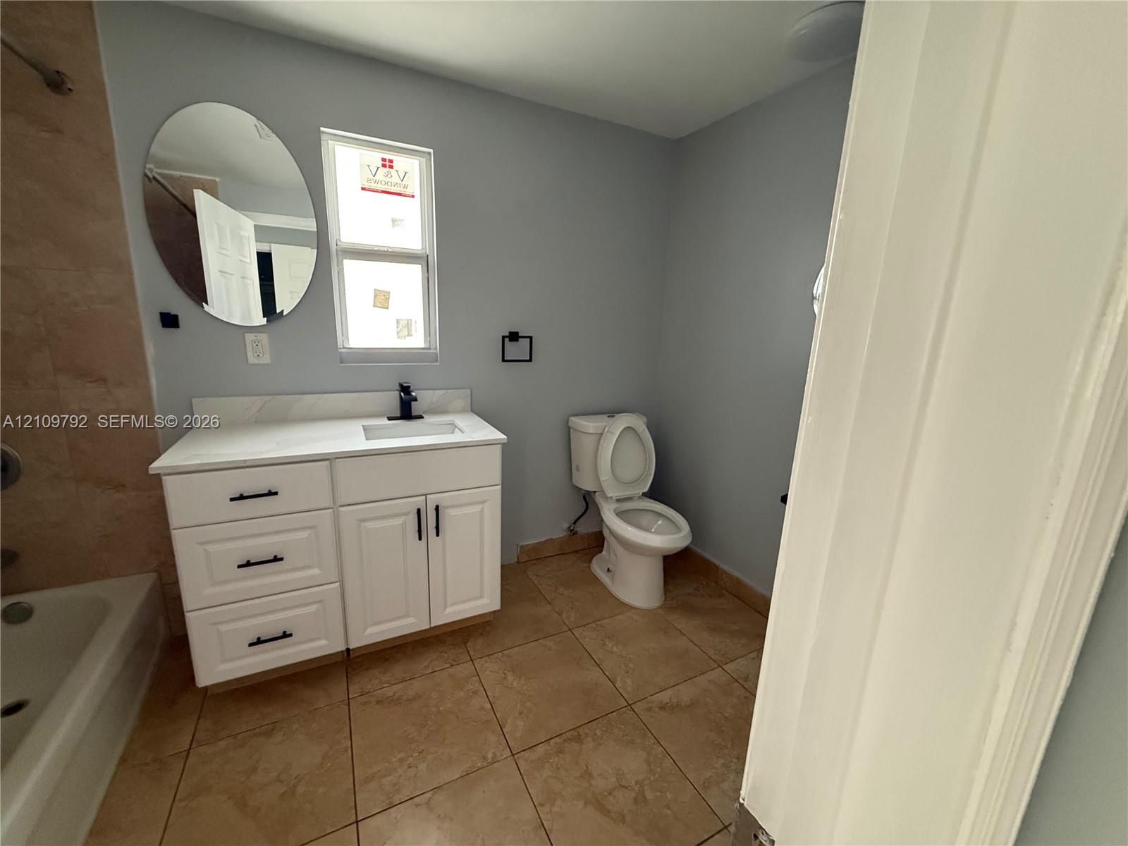 White vanity featuring a stone-look countertop, integrated rectangular sink, matte black faucet, and oval mirror, complemented by tile flooring and tiled tub surround