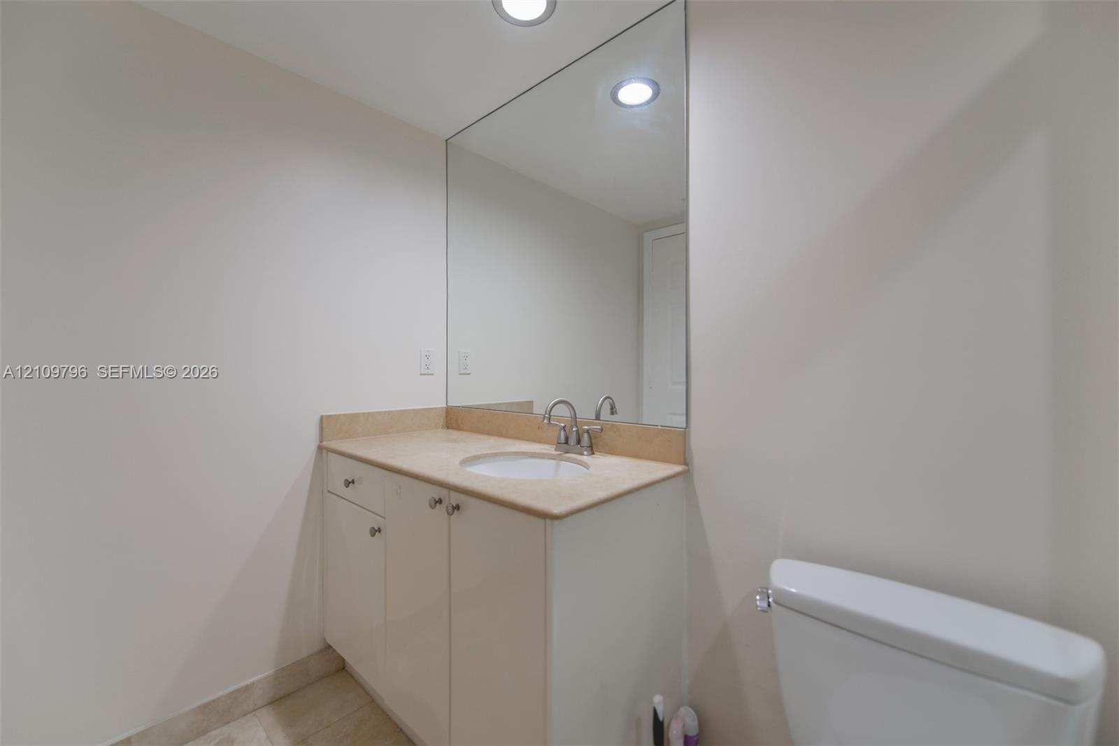 Bathroom vanity featuring a stone-surface countertop, an integrated undermount sink, brushed-metal hardware, a large frameless wall-mounted mirror, and recessed ceiling lighting Bathroom vanity featuring a stone-surface countertop, an integrated undermount sink, brushed-metal hardware, a large frameless wall-mounted mirror, and recessed ceiling lighting
