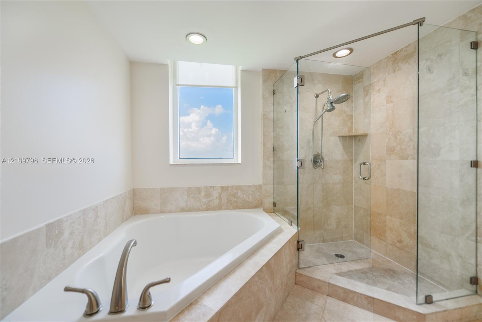 Corner soaking tub with stainless steel faucet, frameless glass-enclosed shower featuring natural stone wall tile, built-in recessed stone shelving, and recessed lighting