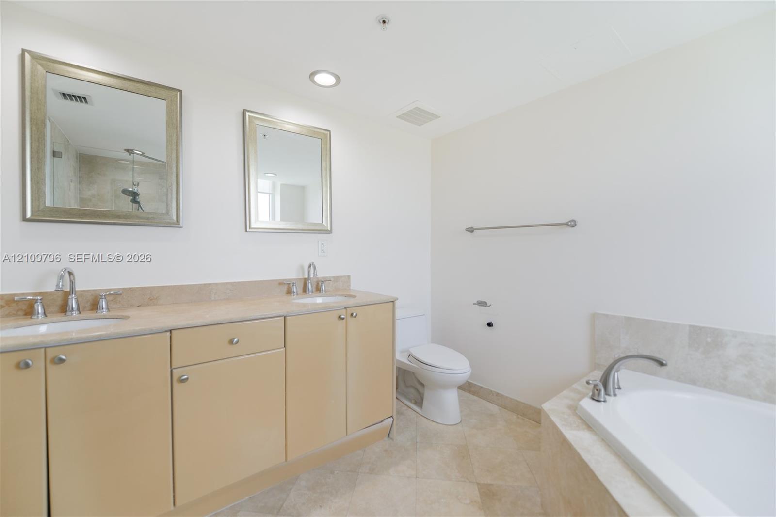 Dual-sink vanity with neutral-toned cabinetry, light stone-finish countertop, two framed wall mirrors, stone tile flooring, and a drop-in soaking tub with a stone tile surround
