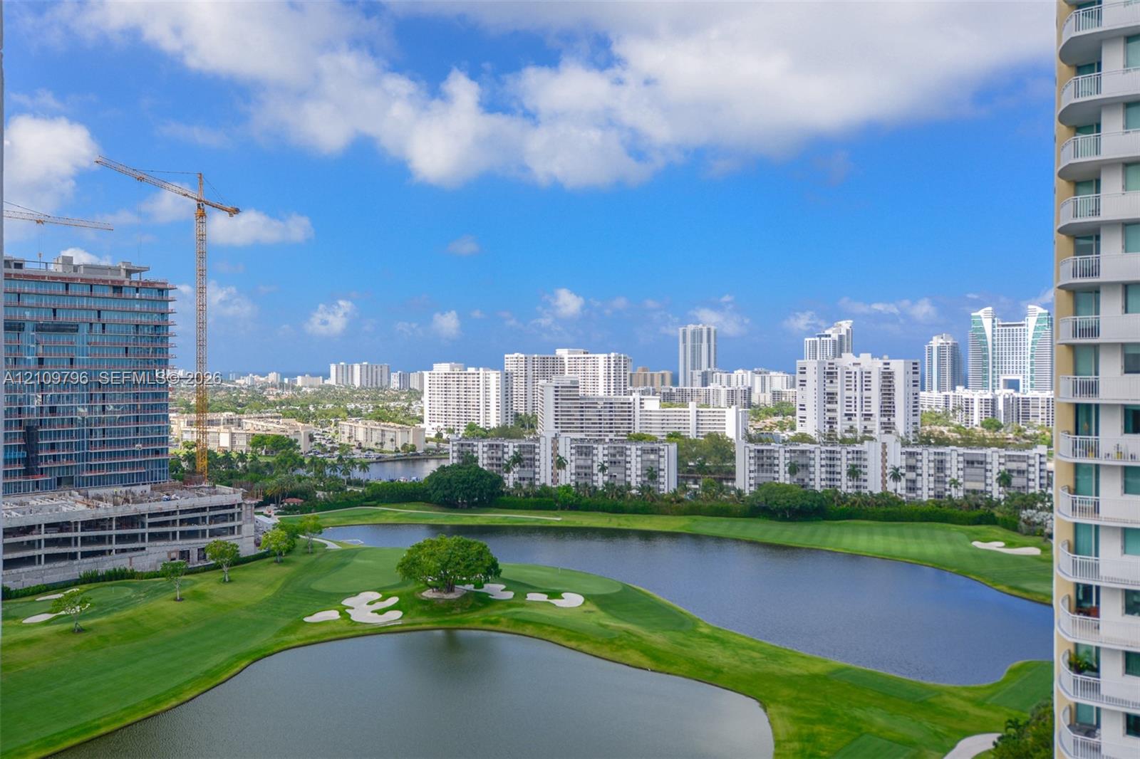 High-rise balcony featuring glass railings, overlooking a sprawling landscape with a manicured golf course, multiple water features, and expansive urban skyline