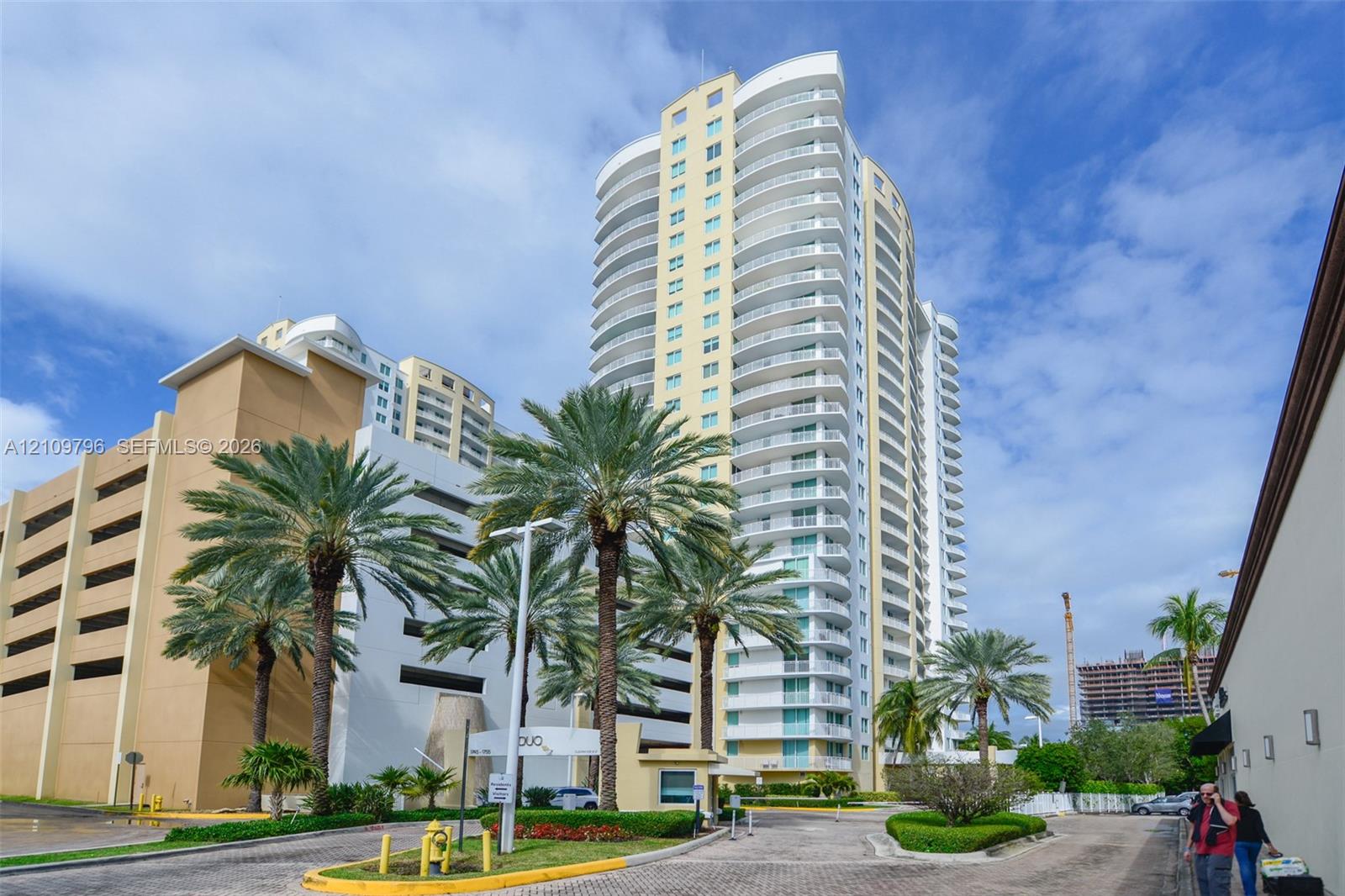 High-rise residential tower featuring curved balconies, floor-to-ceiling glass windows, and an attached multi-level parking structure, situated within landscaped grounds containing mature palm trees